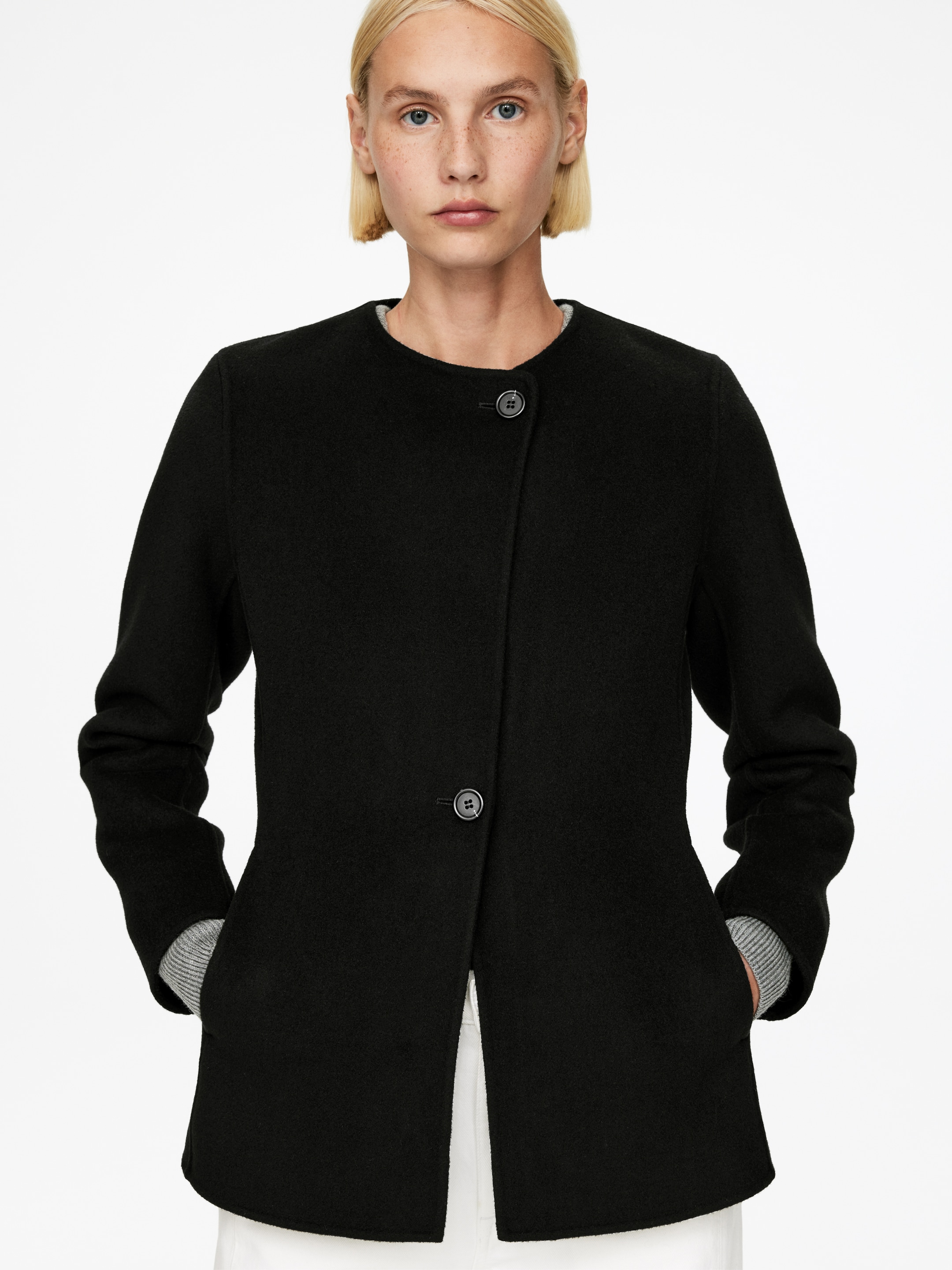 Agrandir l'image: Le produit est un manteau noir sans col, avec une finition texturée et un col rond. Il présente une fermeture à boutons décentrée avec deux boutons sombres, et les mains reposent dans ses poches latérales, révélant les poignets côtelés gris d'un sous-vêtement.