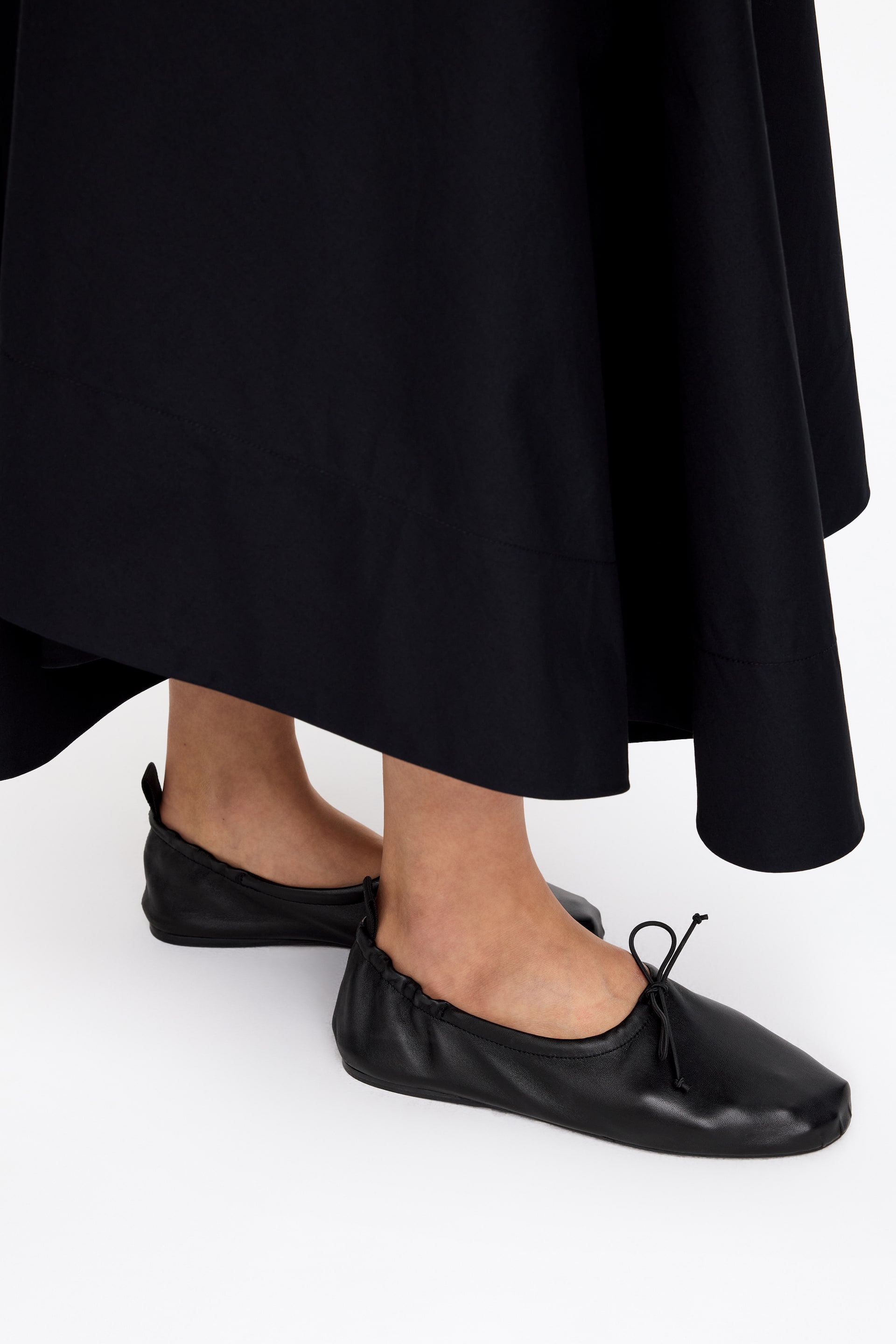 Grösseres Bild ansehen: Schwarze Ballerinas aus Leder werden getragen und zeigen leicht nach rechts. Sie verfügen über eine quadratische Spitze, einen elastischen Einstieg und eine Schleife mit Kordelzug, die oben gebunden ist. Eine kleine Zuglasche ist an der Ferse sichtbar.