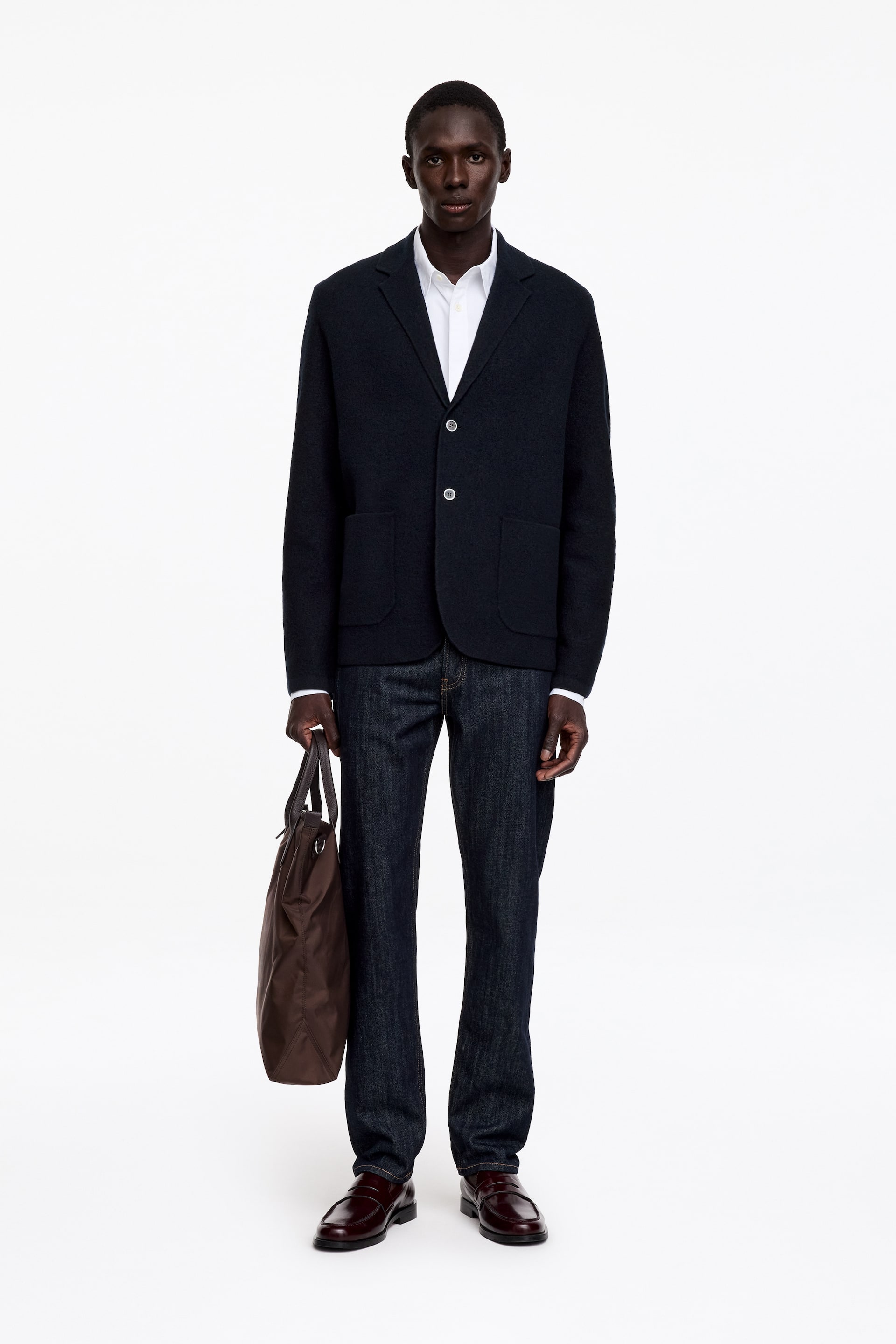 View larger image: A dark navy knitted blazer featuring a two-button fastening and large patch pockets is being worn by the model over a white collared shirt and dark jeans. The model is standing and holding a brown tote bag in one hand.