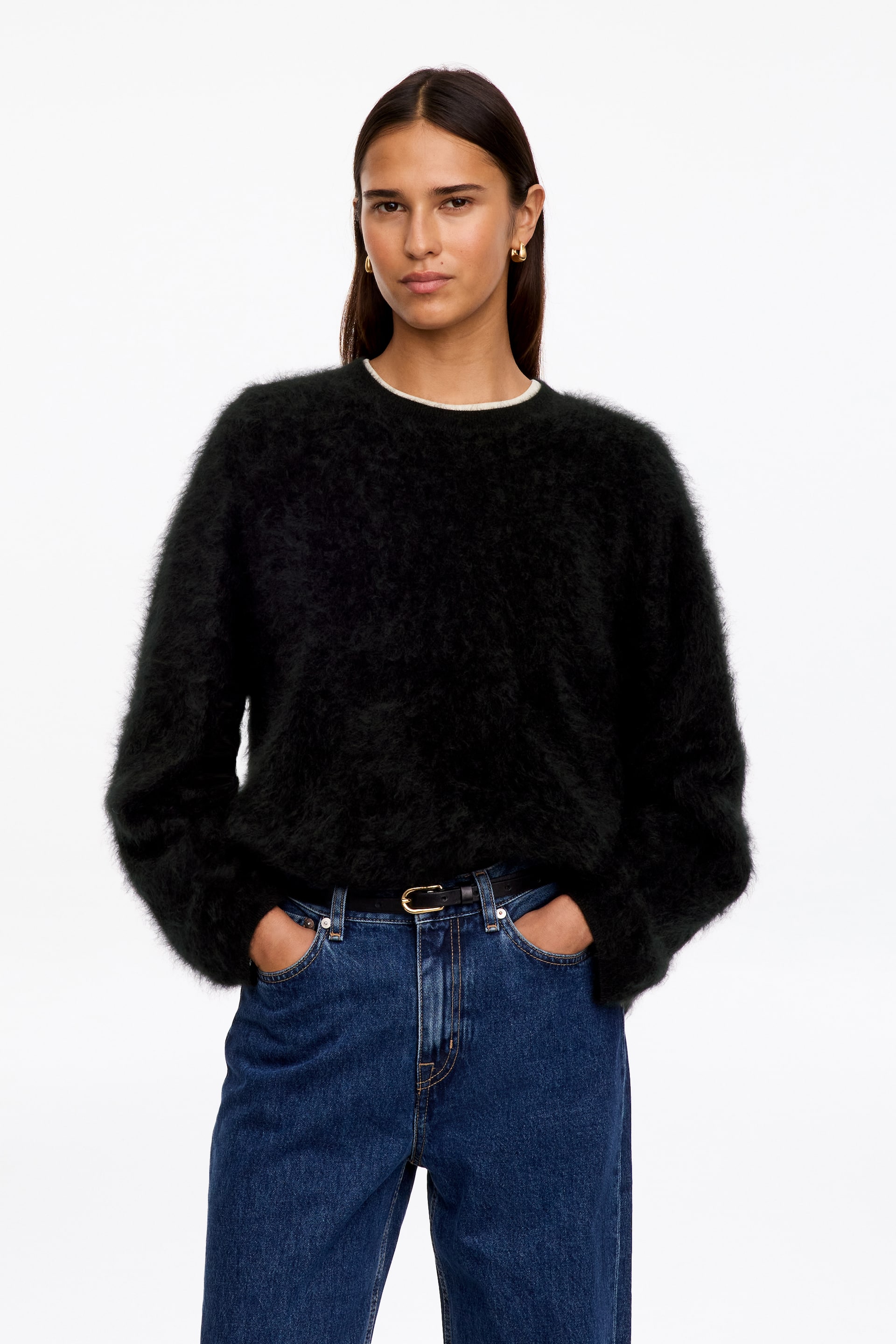 Agrandir l'image: Un pull noir à col rond, à la texture douce et brossée, est porté par le mannequin. Ce pull à coupe décontractée présente des épaules tombantes et est rentré dans un jean bleu avec une ceinture noire, laissant apparaître un fin col blanc en dessous.