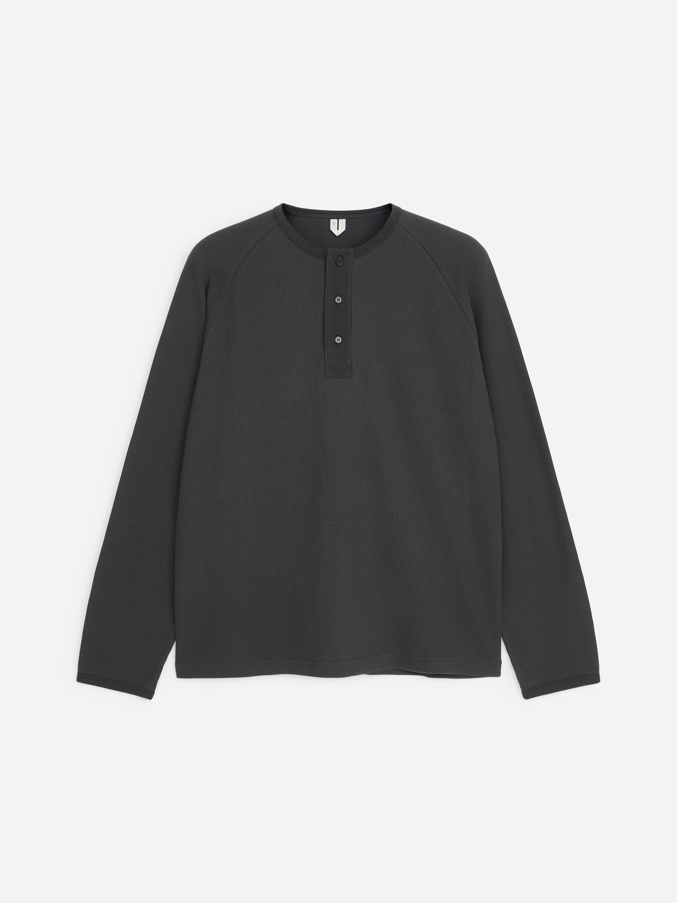 Una camisa Henley de manga larga en gris oscuro, con textura tipo gofre, mangas raglÃ¡n y tapeta parcial con botones, llevada por el modelo. La camisa se combina con unos pantalones de color gris oscuro, y el modelo tiene las manos metidas en los bolsillos.
