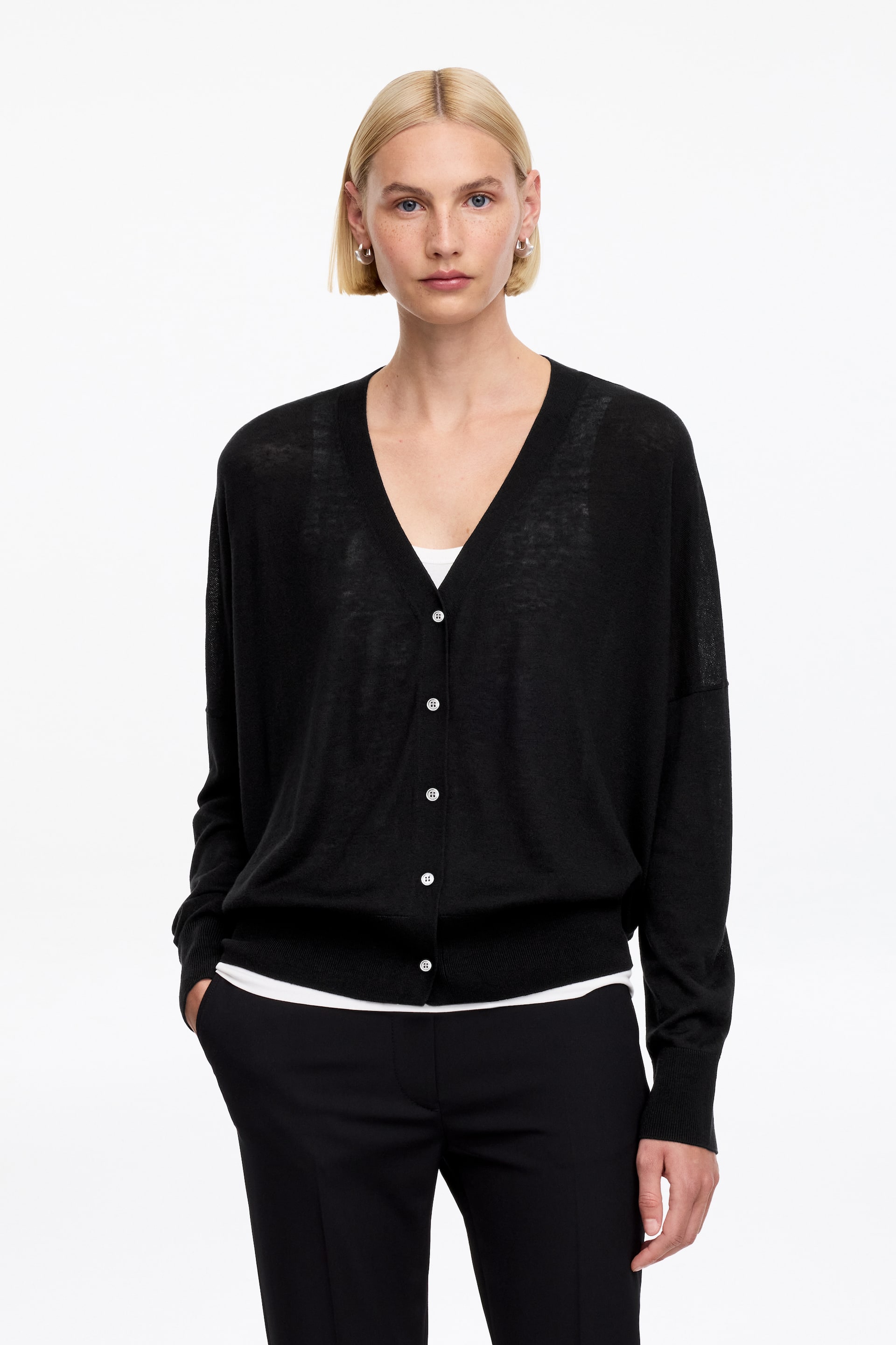 Visa större bild: En svart kofta vänd framåt, med en genomskinlig, finstickad konstruktion med en v-ringning och en knäppning framtill, med vita knappar längs framsidan. Den är designad med långa, lösa ärmar som avslutas med ribbade muddar och en ribbad fåll, och kan bäras över en vit topp.