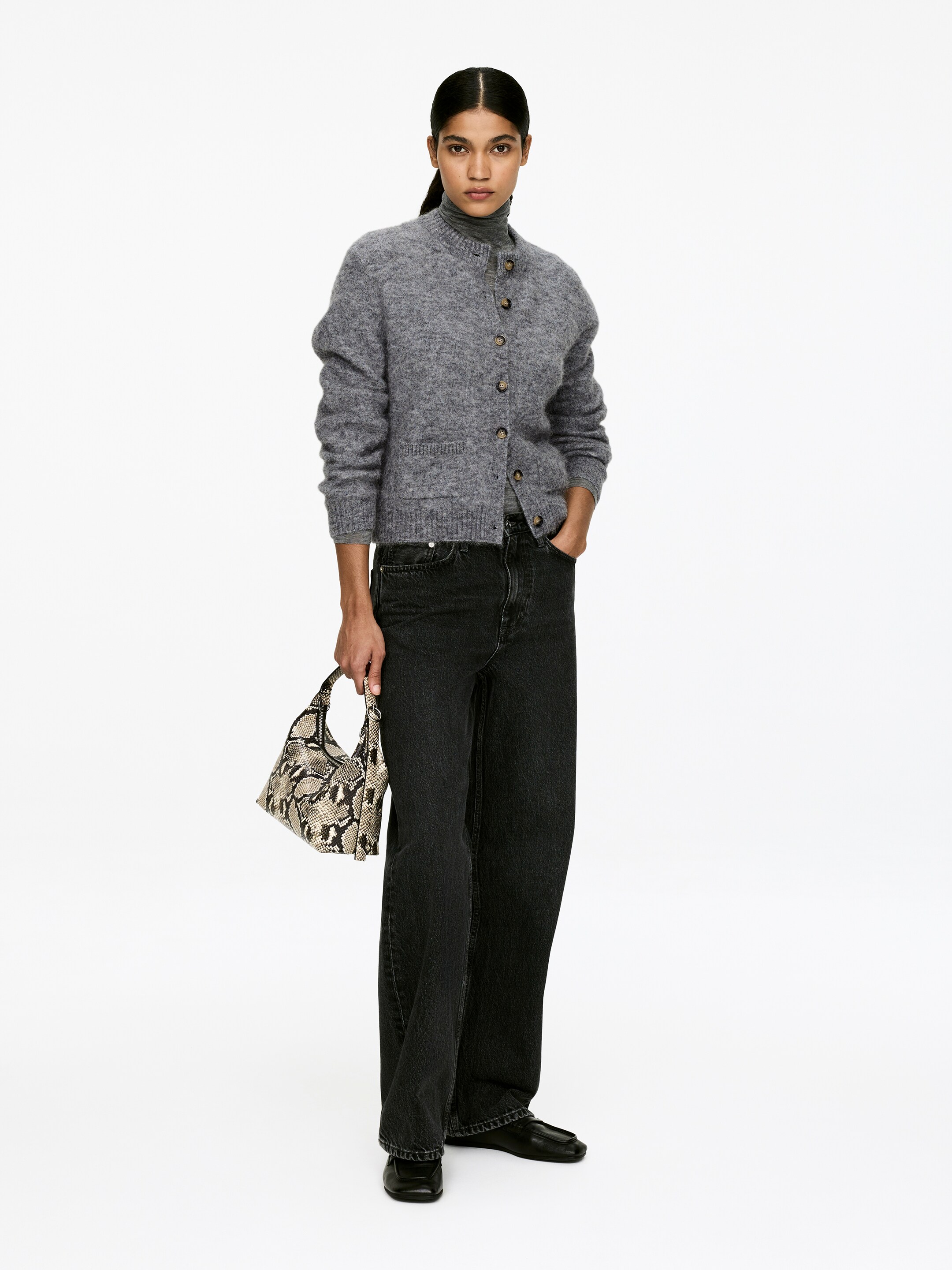 Agrandir l'image: Un cardigan en tricot gris mélange est présenté de face, avec un col rond, une fermeture à boutons sur le devant avec des boutons marron, et deux poches plaquées à l'avant. Il est stylé sur un col roulé gris et associé à un jean noir à jambes larges, avec un sac à bandoulière à motif peau de serpent tenu en main.