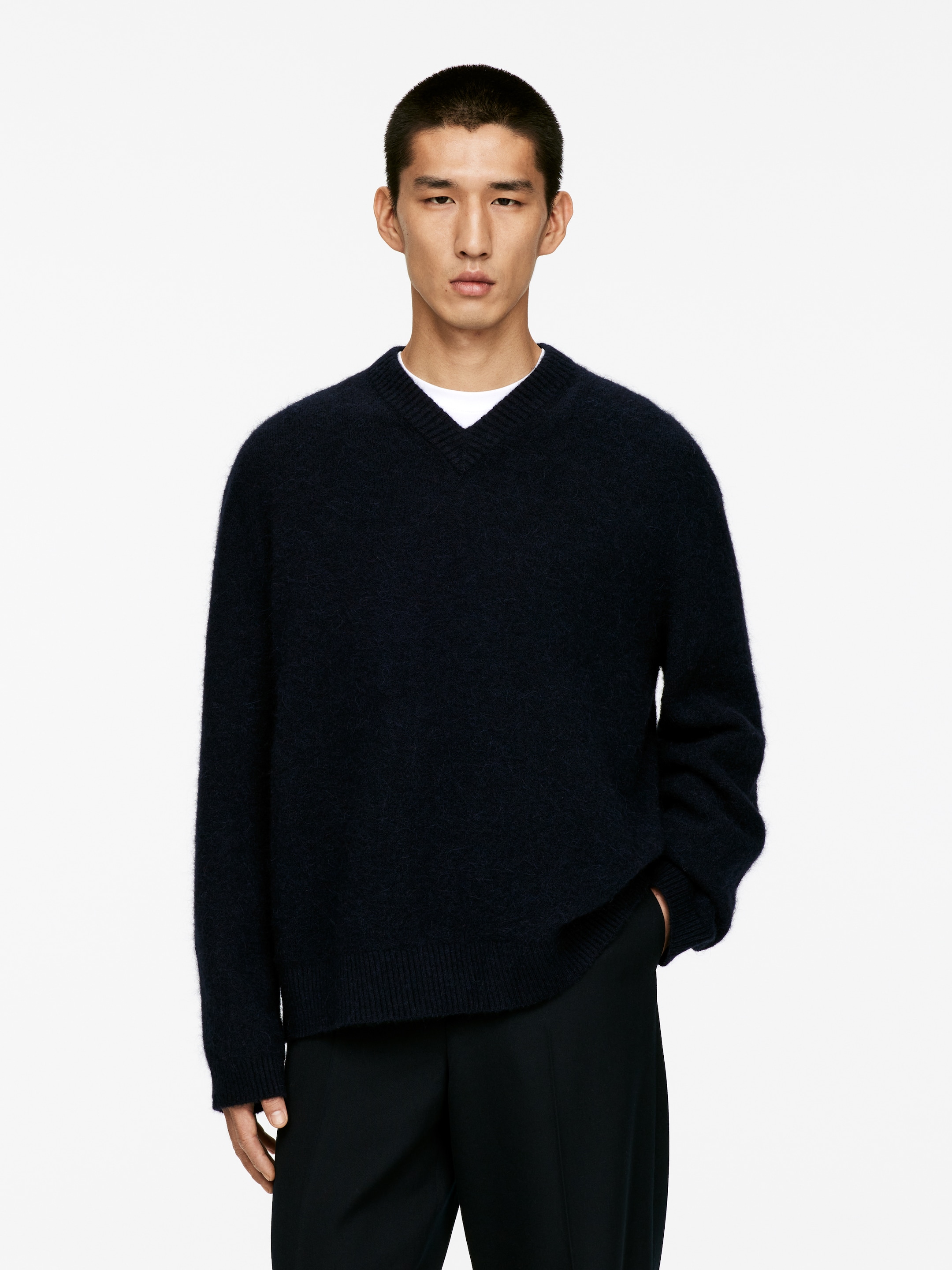 Ingrandisci l'immagine: Un maglione blu scuro a V è presentato su un modello rivolto in avanti, caratterizzato da una texture pelosa, polsini a coste e un orlo a coste. Una t-shirt bianca con scollo rotondo è visibile sotto il maglione a V.