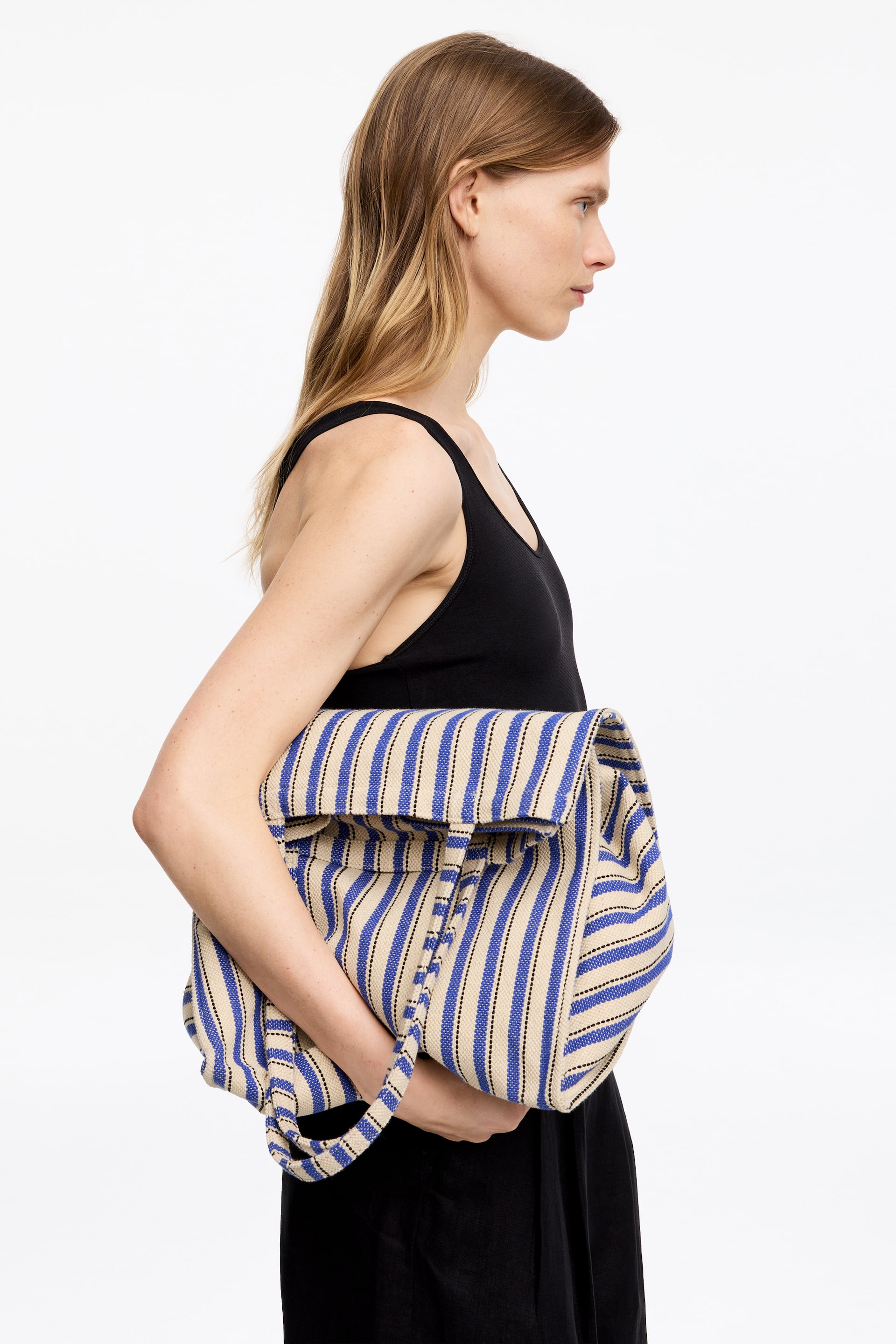 Se større billede: En rummelig tote-taske i bomuldslærred med et mønster af blå, beige og sorte striber, bæres af modellen. Hun holder i en af dens håndtag, mens hun har tasken under armen.