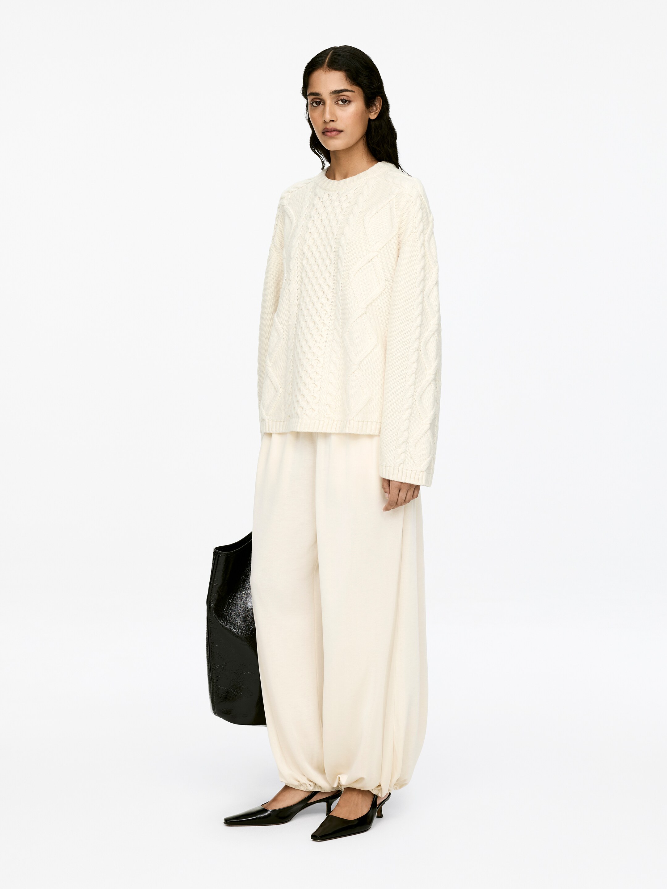 View larger image: A cream cable knit jumper, featuring a crew-neck, long sleeves, and ribbed trims, is being worn by the model. The jumper displays a relaxed fit with intricate cable patterns.