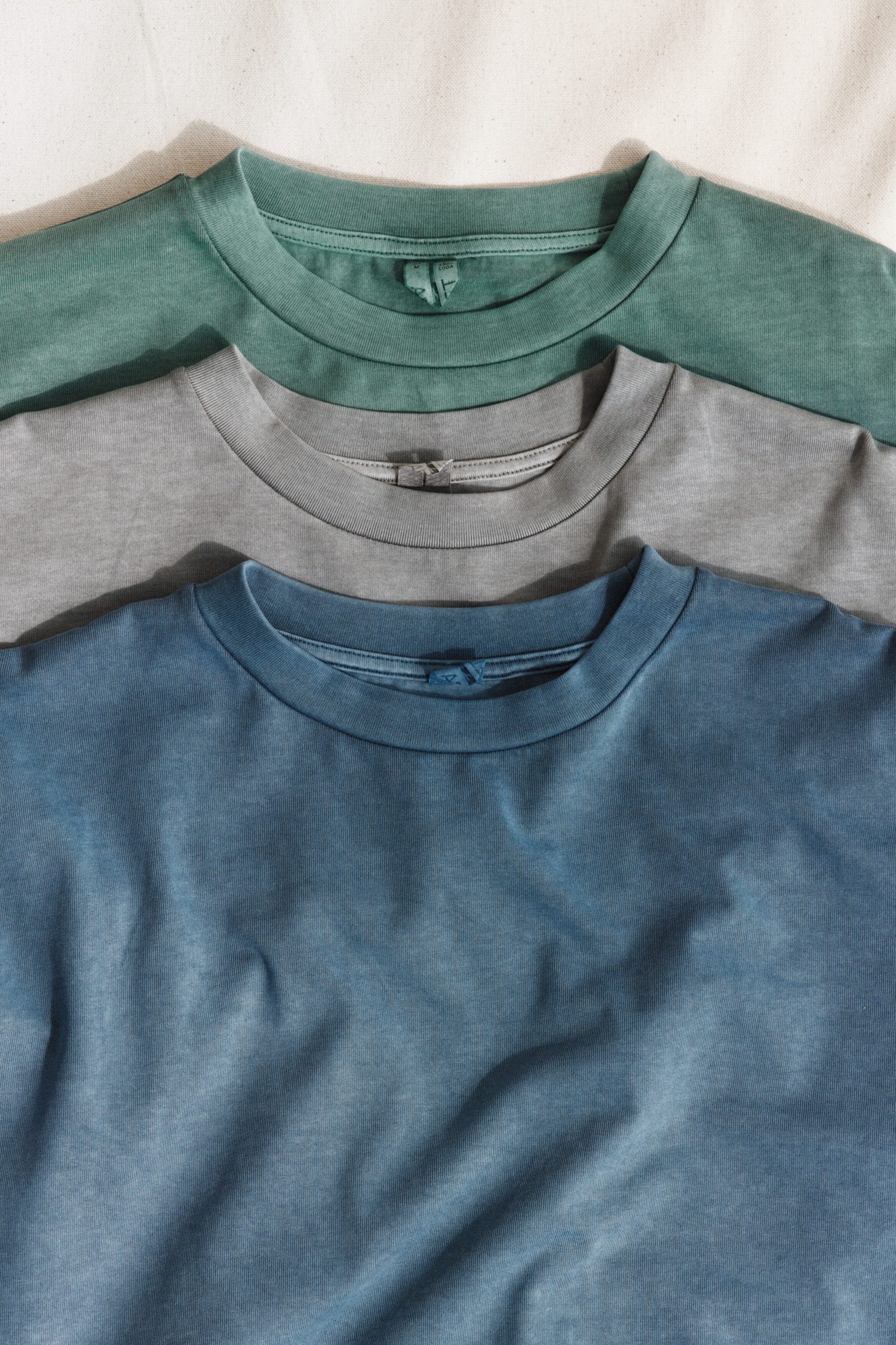 Agrandir l'image: Trois t-shirts sont superposés horizontalement, montrant un t-shirt vert pâle sur le dessus, un t-shirt gris clair au milieu et un t-shirt bleu délavé en bas, tous dotés de cols ronds, de zones supérieures de la poitrine, d'une texture de tissu douce et lavée, et de cols ras-du-cou côtelés.