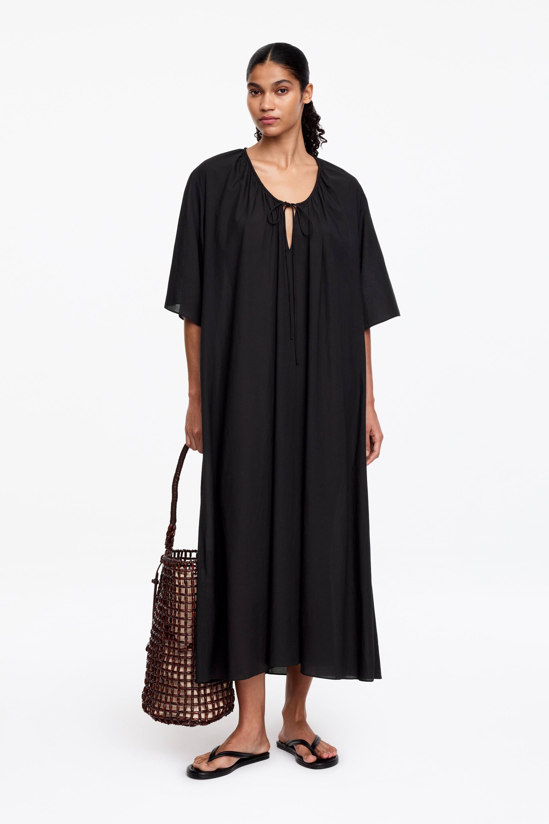 Ver imagem maior: Um vestido maxi preto é apresentado de frente, com um decote redondo franzido com uma abertura em V e fecho com laço. É desenhado com mangas curtas e largas e uma silhueta solta, estendendo-se até abaixo do joelho.