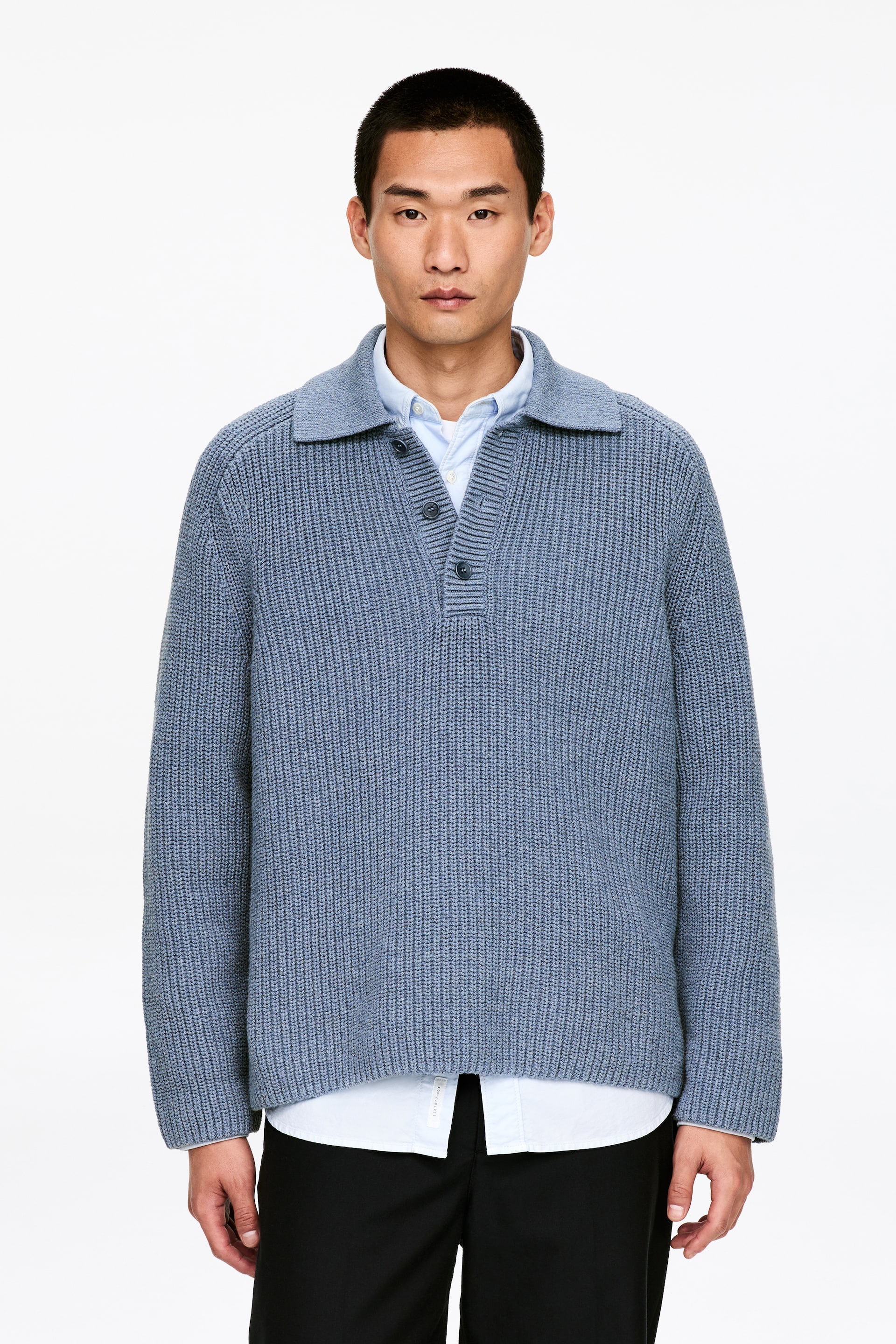Visualizza immagine più grande: Un maglione a coste blu con scollo a polo ampio e abbottonatura a tre bottoni, presenta una vestibilità rilassata ed è stratificato su una camicia azzurra con colletto visibile.