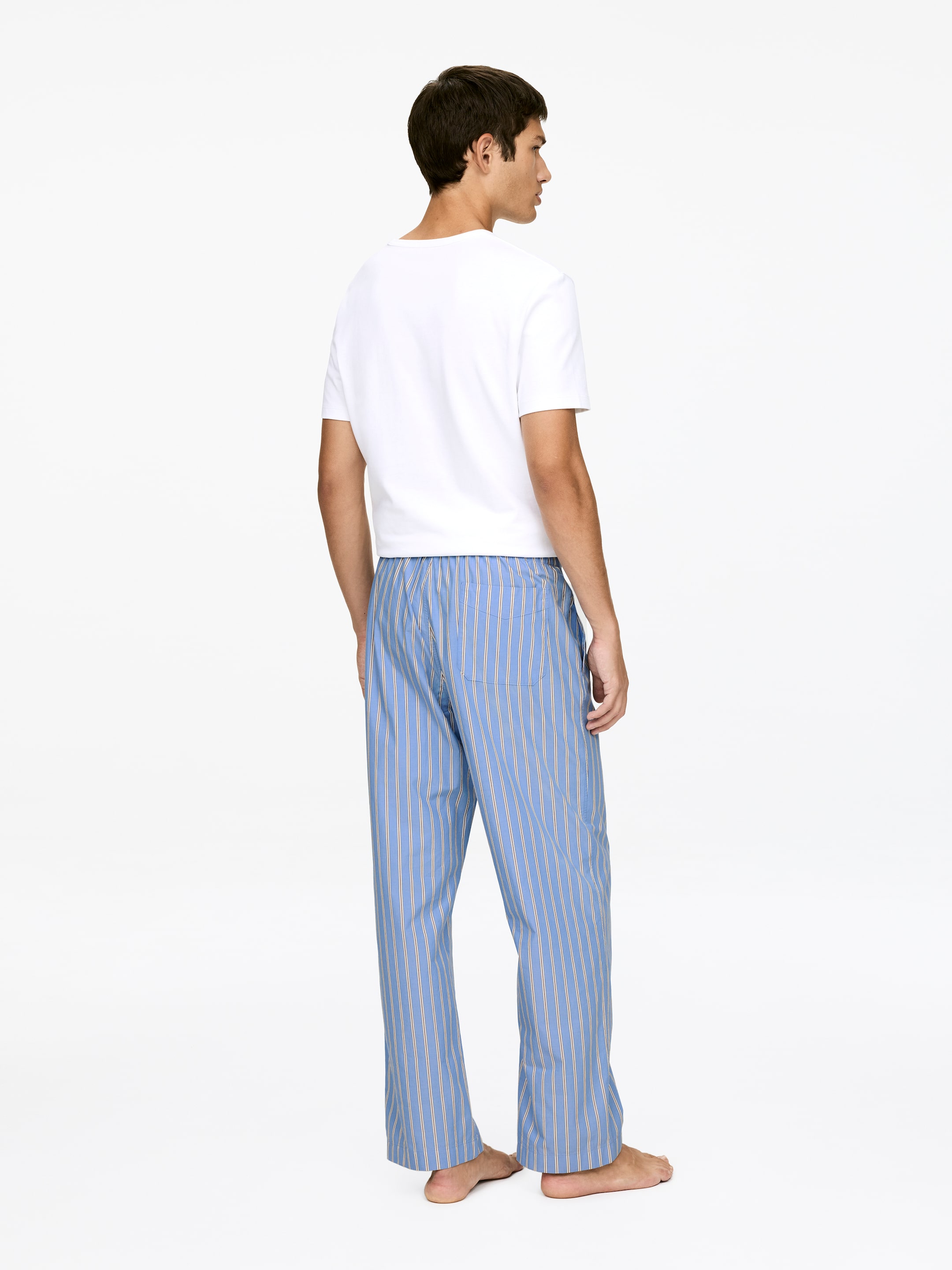 Visualizza immagine più grande: Una maglietta bianca a maniche corte viene indossata con pantaloni del pigiama blu, presentati da un punto di vista posteriore. I pantaloni del pigiama presentano righe verticali bianche e giallo chiaro, una vestibilità rilassata, una vita con elastico e una sola tasca posteriore.