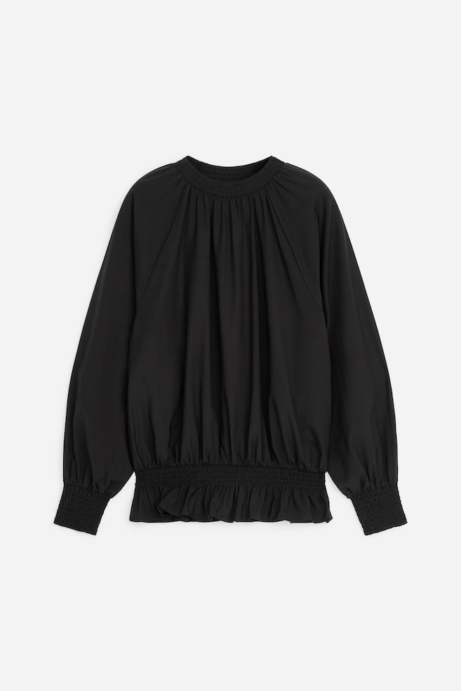 Een zwarte blouse met lange mouwen wordt van voren getoond, met een hoge, gesmokte halslijn en een brede, gesmokte tailleband die een geplooid blouson-silhouet met een gerafelde zoom creëert. De blouse is ontworpen met volumineuze mouwen die eindigen in bijpassende brede, gesmokte manchetten. - 1