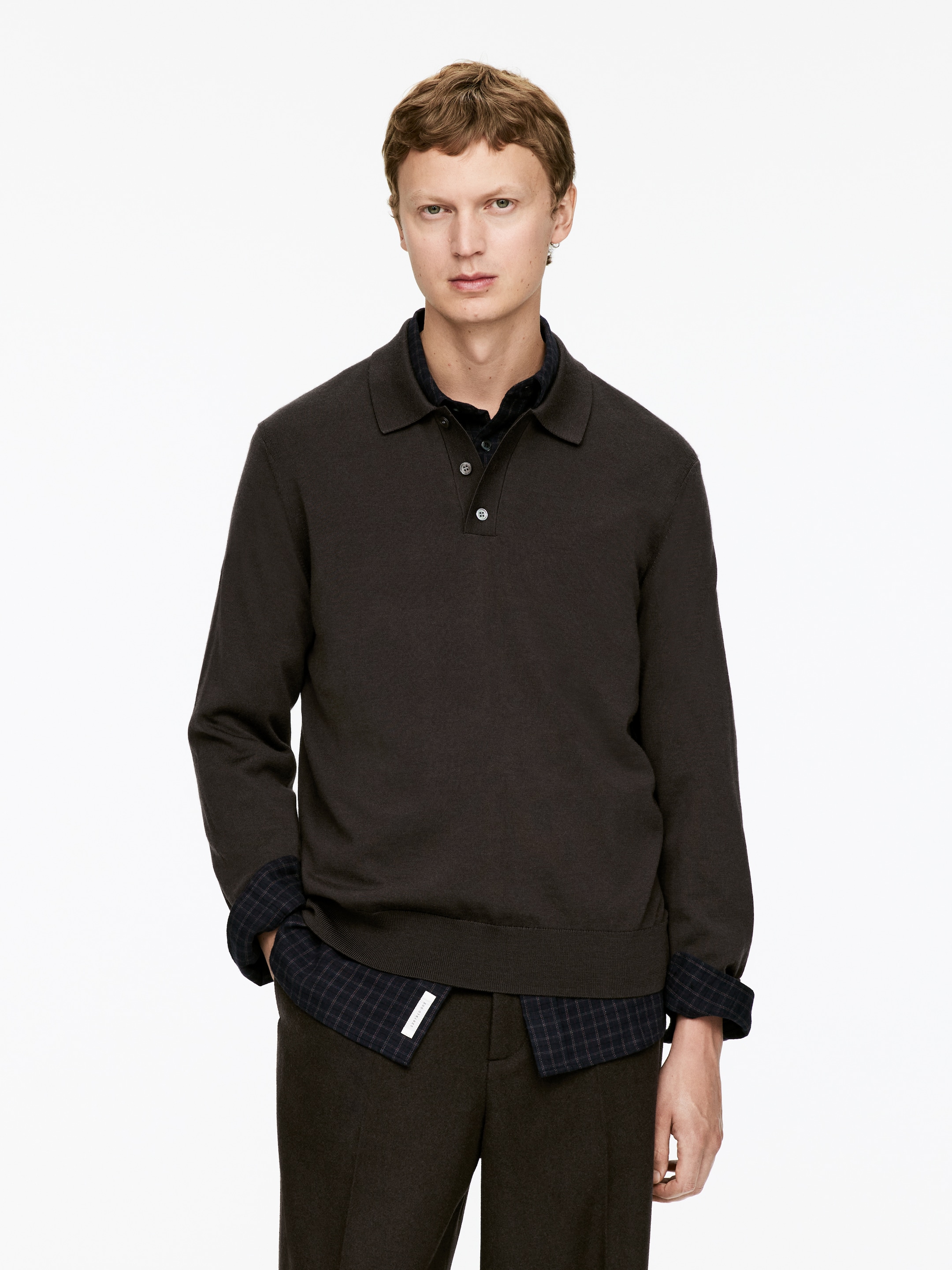 Agrandir l'image: Un polo en tricot marron foncé est porté, face à l'avant. Il présente un col pointu, une patte de boutonnage à trois boutons avec des boutons blanc cassé, et des manches longues avec des poignets et un ourlet bandés. Une chemise à carreaux foncée est superposée en dessous, avec son col, ses poignets roulés et son ourlet visibles.