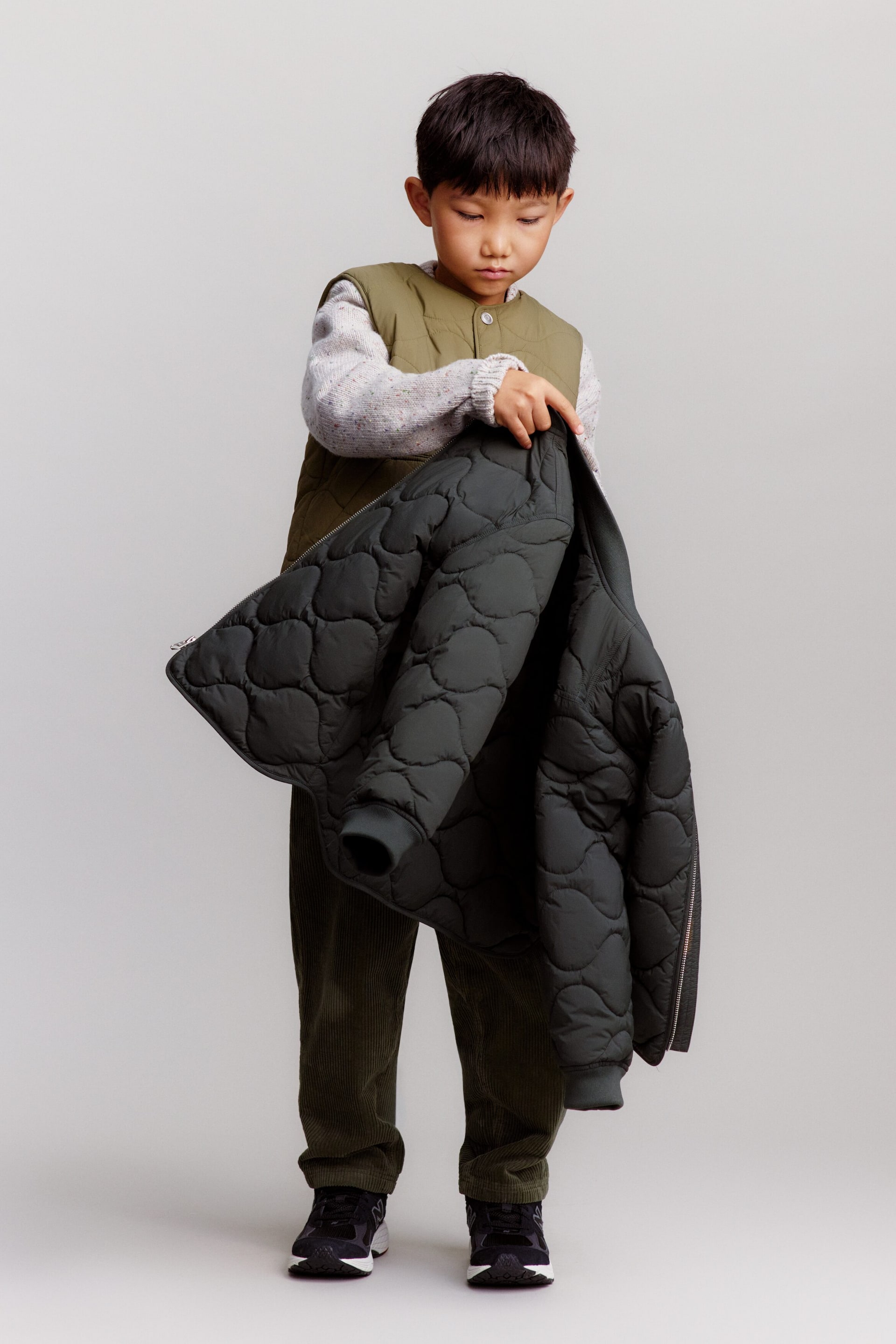 Agrandir l'image: Un enfant se tient de face, portant un pull gris clair superposé sous un gilet matelassé vert olive, associé à un pantalon en velours côtelé vert foncé à coupe décontractée et des baskets noires. L’enfant tient une veste légère matelassée vert foncé avec poignets côtelés, mettant en valeur son matelassage ondulé texturé.