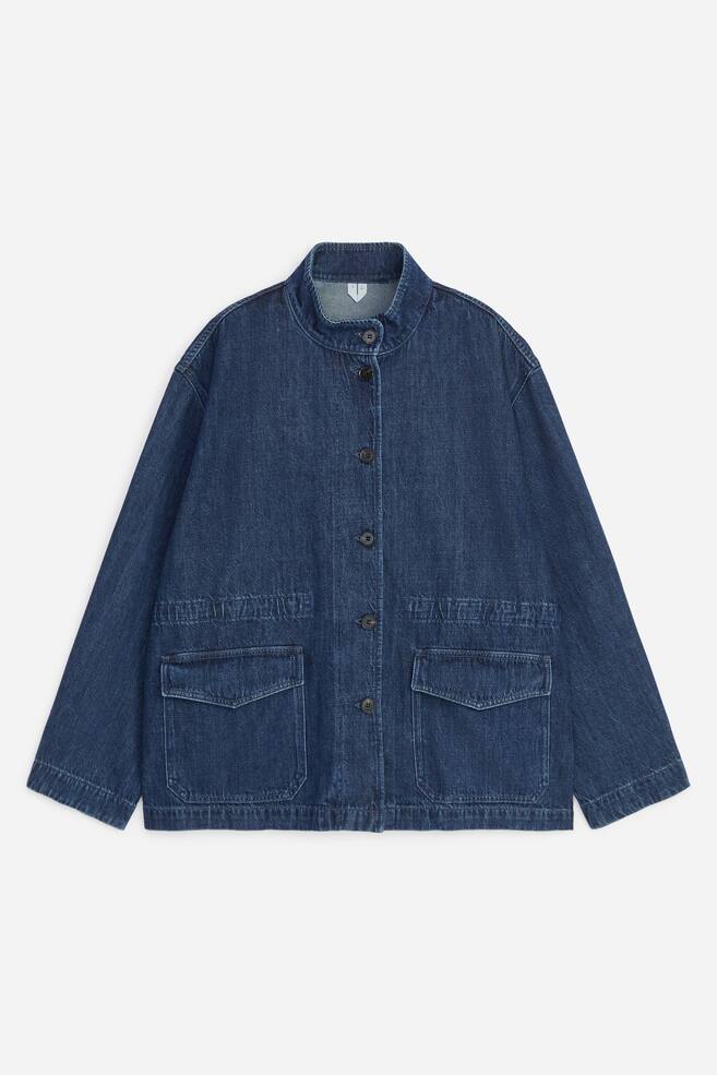 A dark blue denim jacket is shown from the front, featuring a band collar, a central button fastening, and long sleeves with dropped shoulders. It has two large front patch pockets with flaps and gathered elasticated detailing along the waistline. - 1