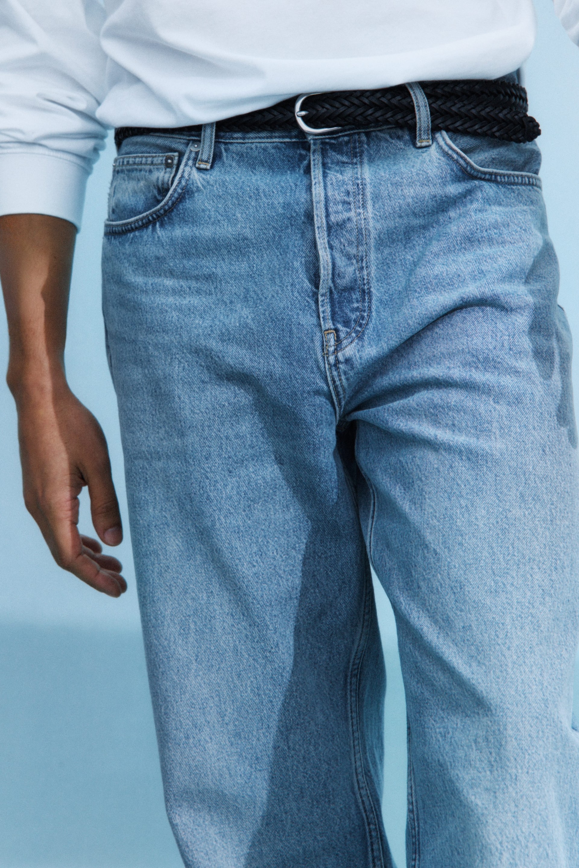 Visualizza l'immagine ingrandita: Jeans in denim blu chiaro con lavaggio leggero presentano una chiusura con bottoni visibili e tasche anteriori. Una cintura intrecciata nera è infilata nei passanti, e una camicia bianca a maniche lunghe è indossata infilata nei jeans.