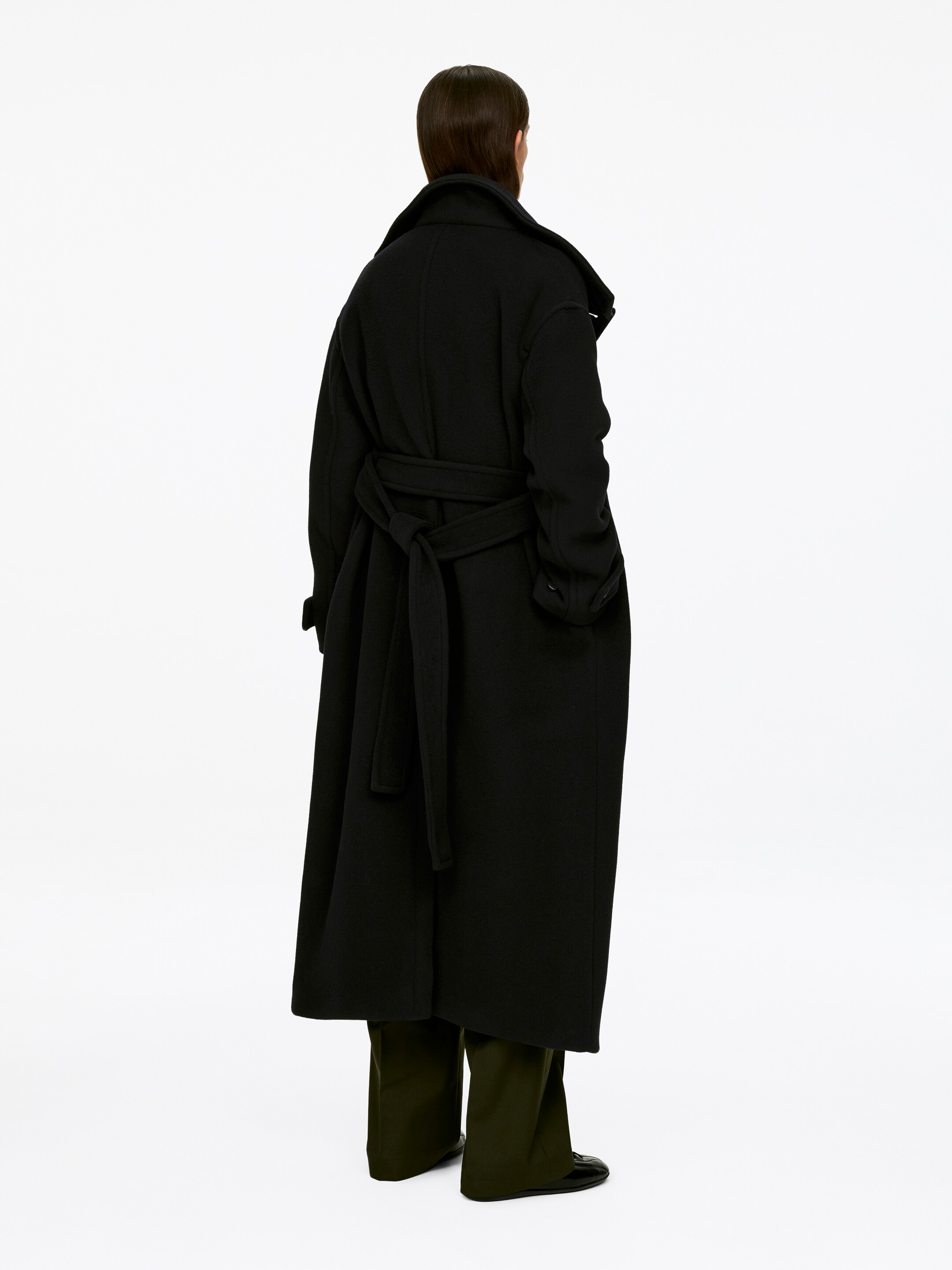 Se større billede: En sort belted maxi frakke bæres af modellen, som vender ryggen til. Den oversized frakke har en opretstående krave og er belted bagpå med en knude. Den lange kant dækker mørkegrønne bukser, og modellens venstre hånd hviler i en sidelomme.