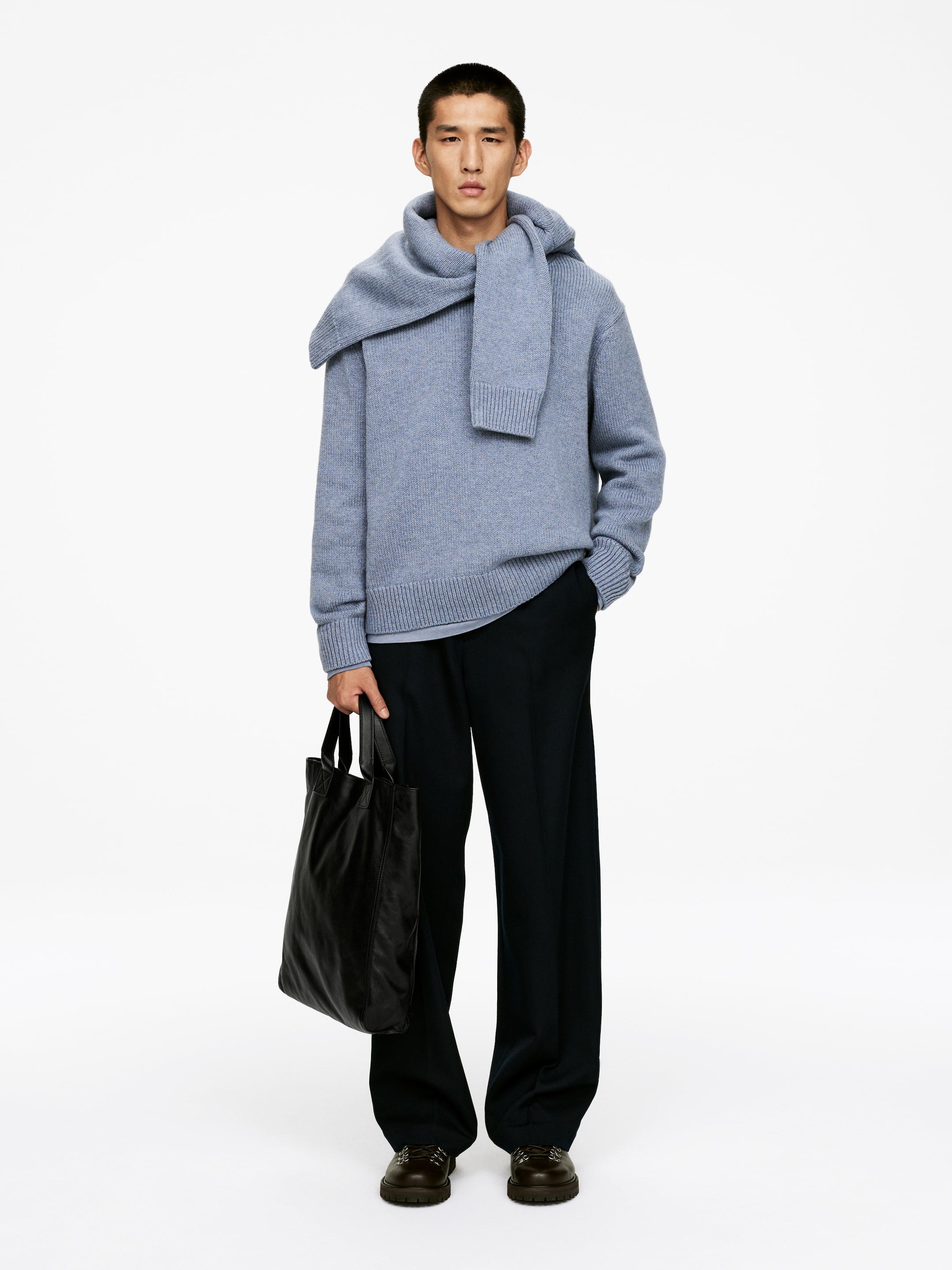 Größeres Bild ansehen: Ein Outfit, das nach vorne gerichtet ist, präsentiert einen hellblauen Strickpullover mit gerippten Bündchen und Saum, kombiniert mit einem passenden breiten Schal, der um den Hals und die Schultern drapiert ist. Dunkle, weit geschnittene Hosen, dunkelbraune Schuhe mit dicker Sohle und eine schwarze, lederähnliche Tragetasche vervollständigen den Look.