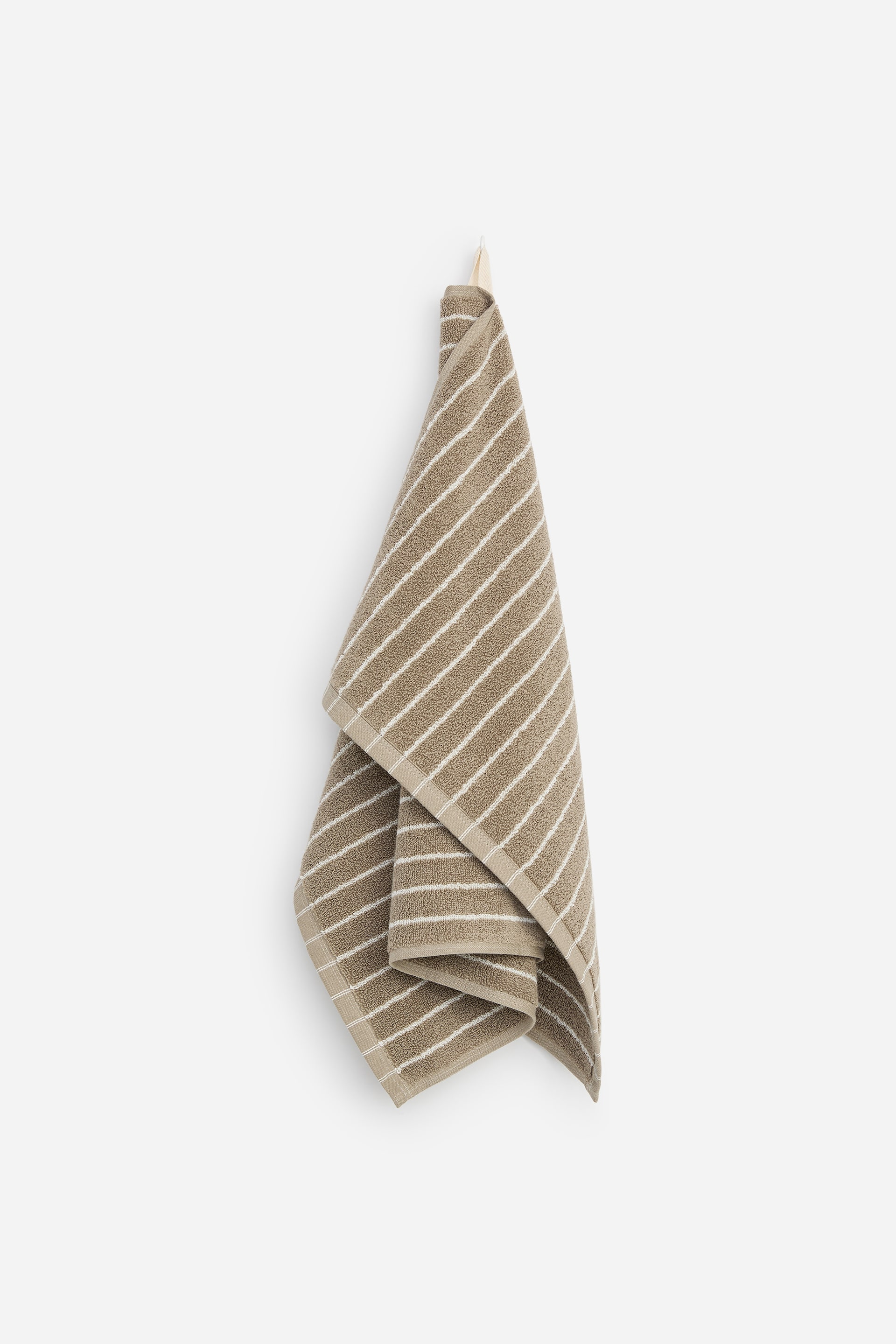 Agrandir l'image: Une serviette à main en éponge de coton beige, ornée de fines rayures blanches horizontales, est suspendue verticalement à un crochet en bois. La serviette tombe en de doux plis superposés, mettant en valeur son tissu texturé et ses bordures beiges unies.