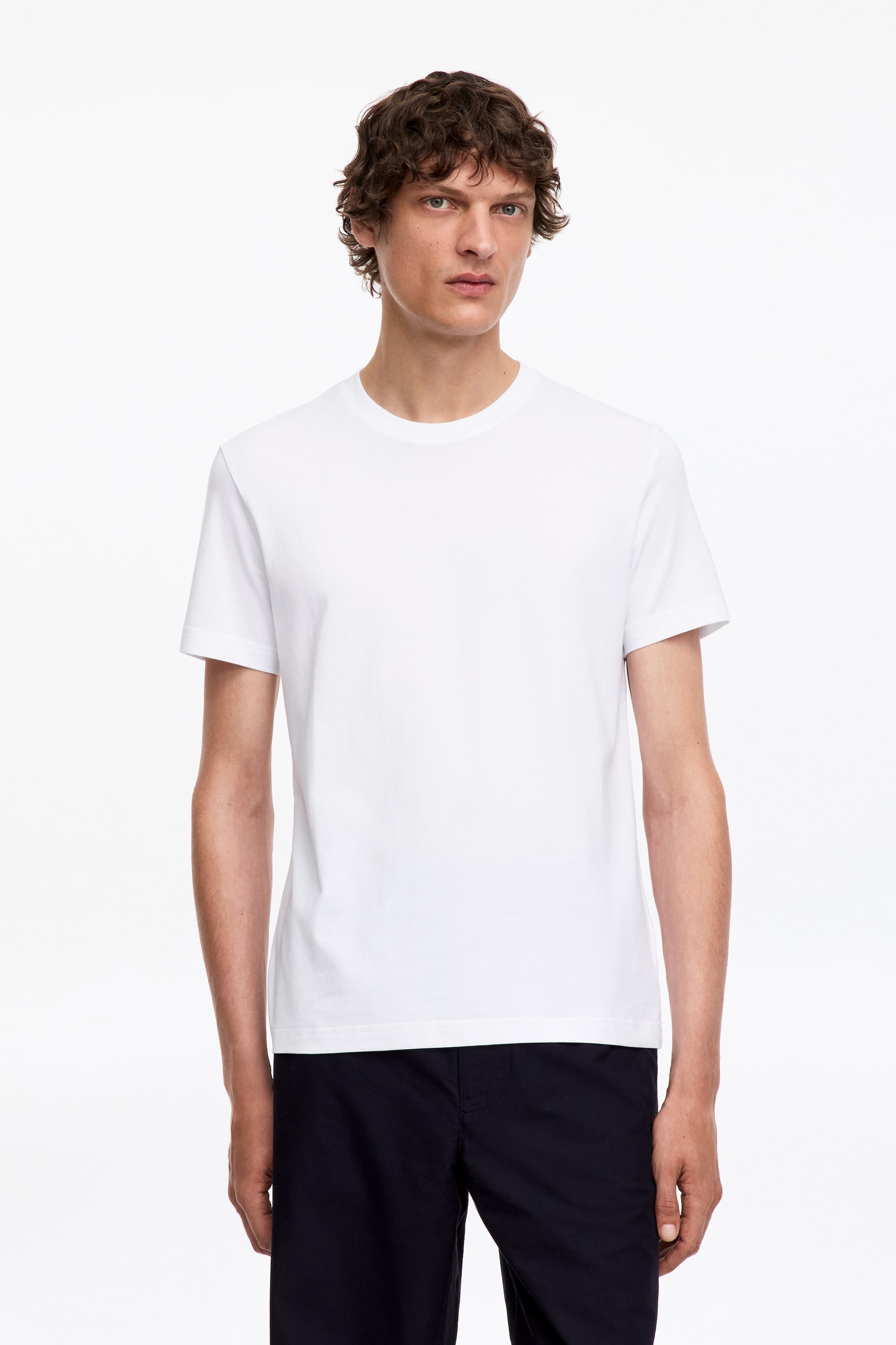 Visa större bild: En vit T-shirt med rund hals bärs av modellen, med en vanlig passform och något kortare, åtsittande ärmar samt en skräddarsydd skärning genom kroppen. Halsringningen är avslutad med 1x1 ribbstickning.