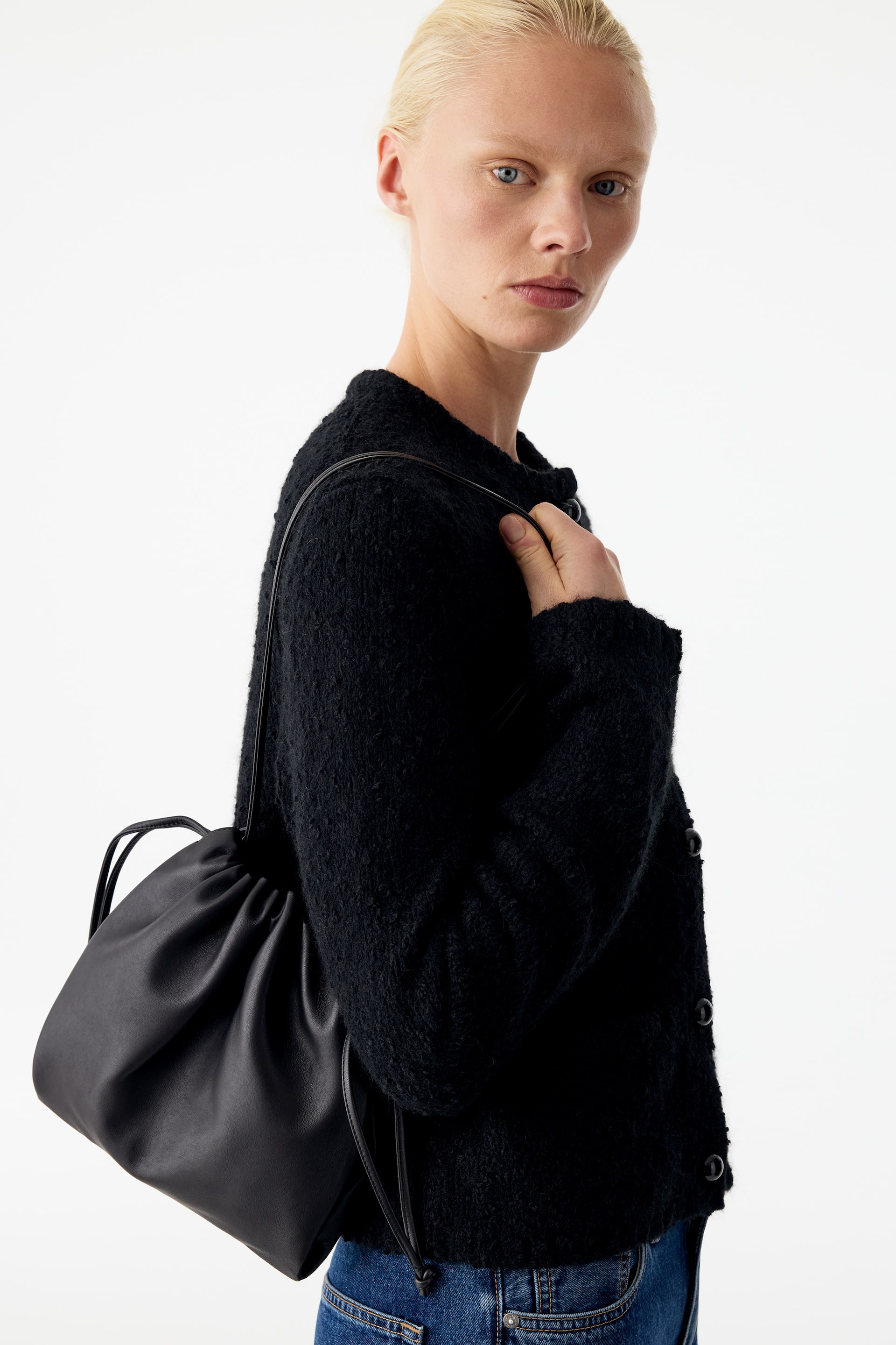 Visa större bild: En svart läderpåse med dragsko och rynkad överdel bärs över axeln, medan modellen håller i dess smala rem. Väskan stylas med en svart cardigan med struktur och blå jeans.
