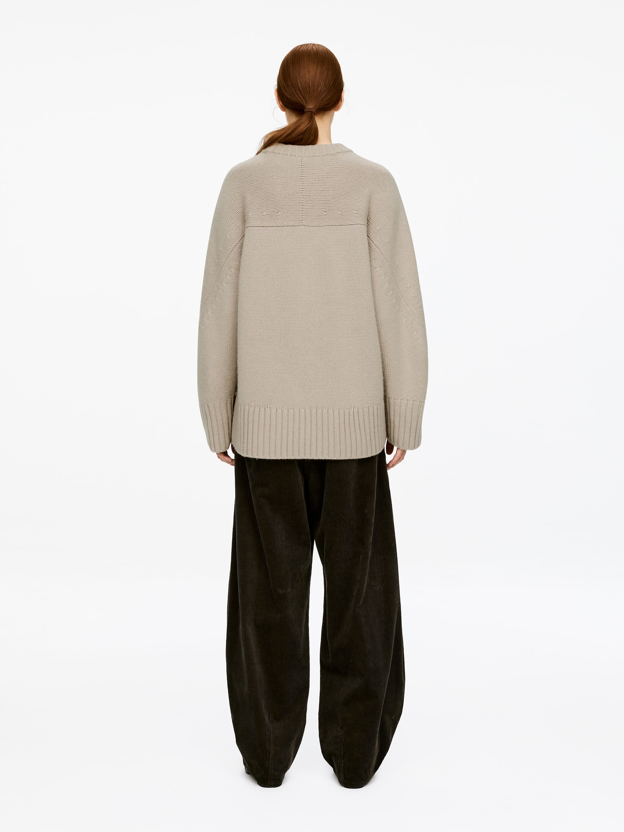 Se større billede: Den Chunky Pure Cashmere Jumper i lysebeige bæres af modellen, set bagfra. Den oversized strik har ribbede manchetter og kant, og en vandret sømdetalje hen over den øvre del af ryggen, stylet med mørkebrune bredbenede fløjlsbukser.