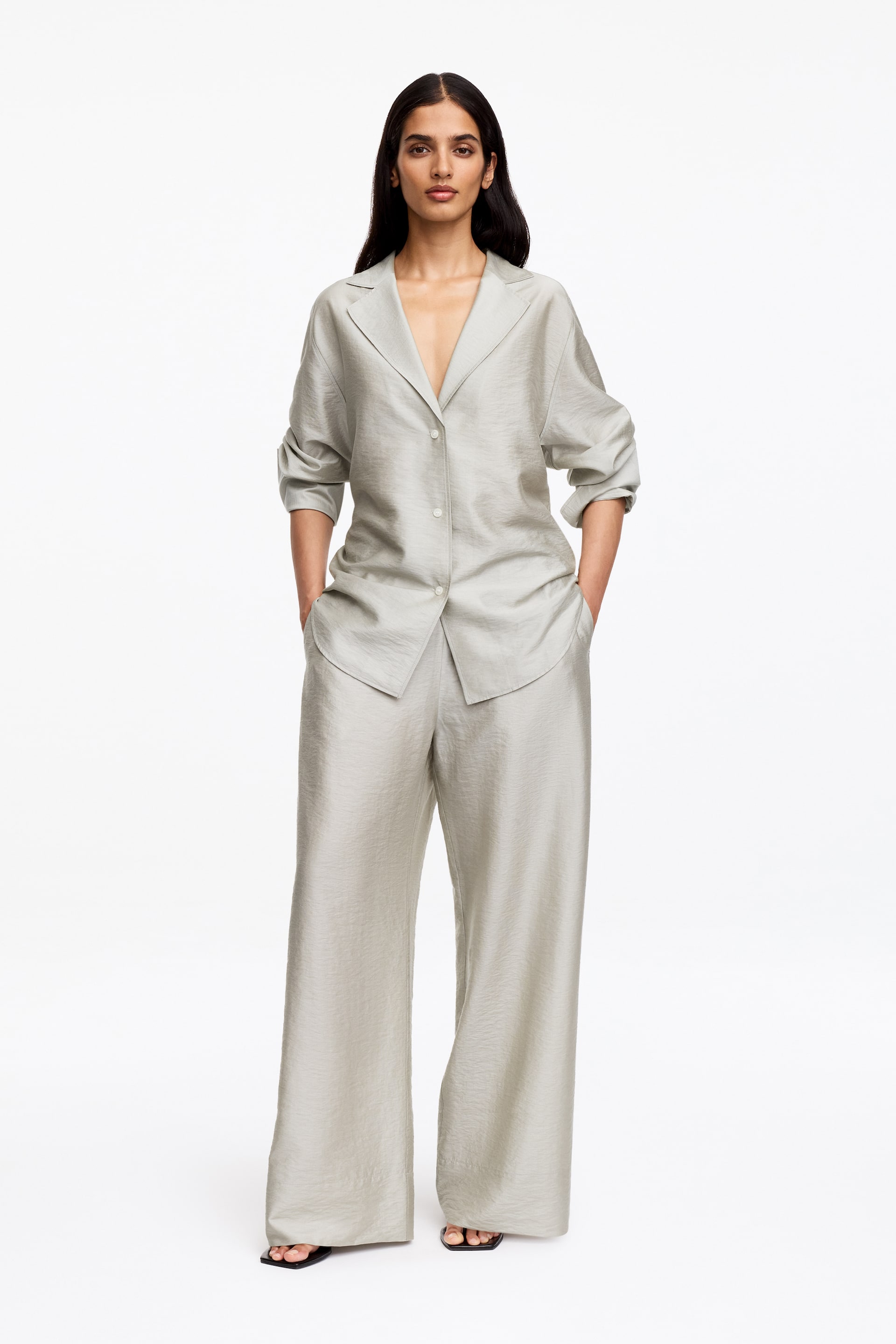 Visualizza immagine più grande: Un set due pezzi scintillante color grigio chiaro è in mostra, composto da una camicia a vestibilità rilassata con colletto a rever, maniche arrotolate e bottoni frontali visibili. I pantaloni abbinati a gamba larga e lunghezza intera presentano una caduta morbida e tasche laterali.