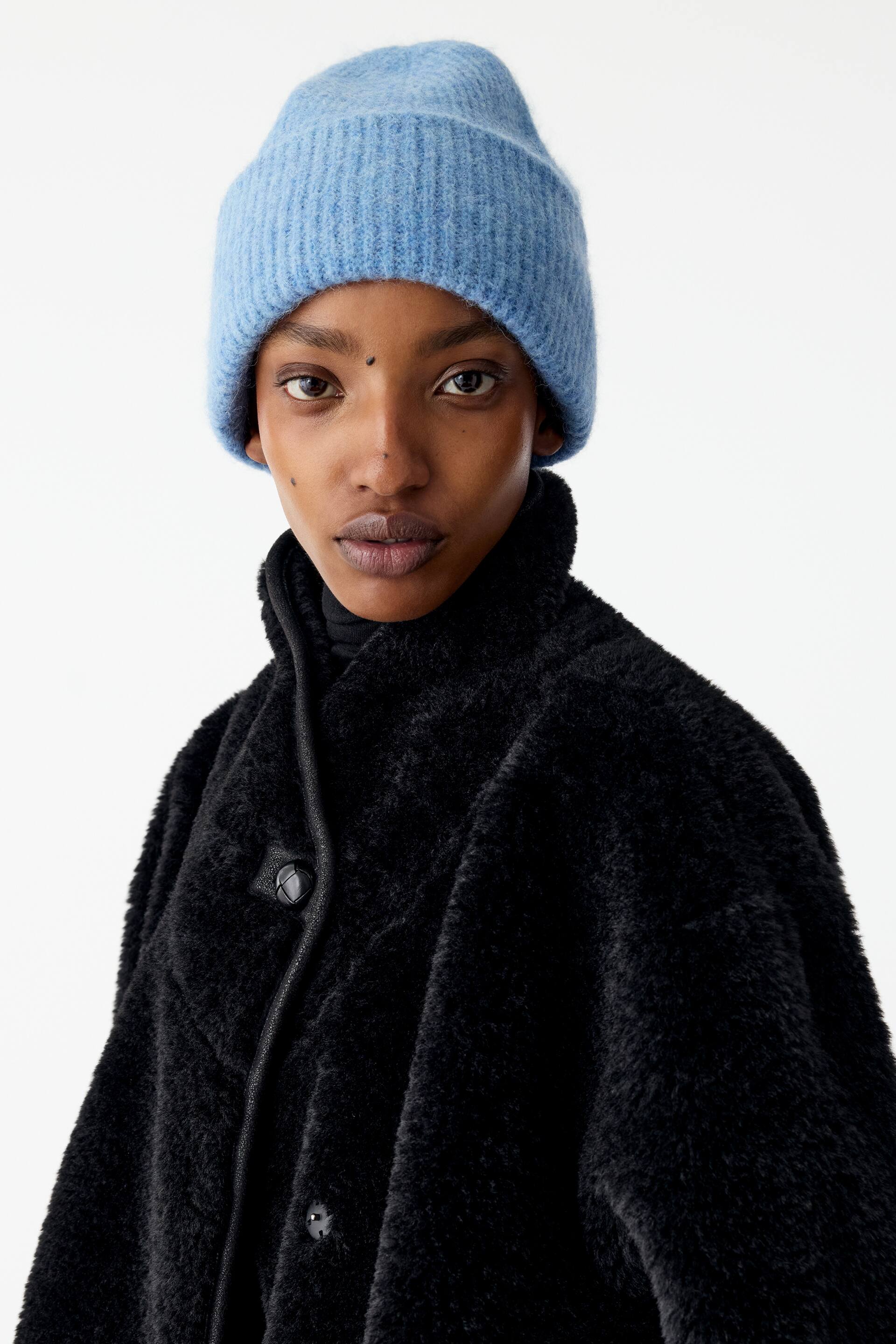 Agrandir l'image: Un bonnet en maille côtelée bleu clair est porté par le mannequin, associé à un manteau noir texturé doté d’un col montant et de fermetures à boutons.
