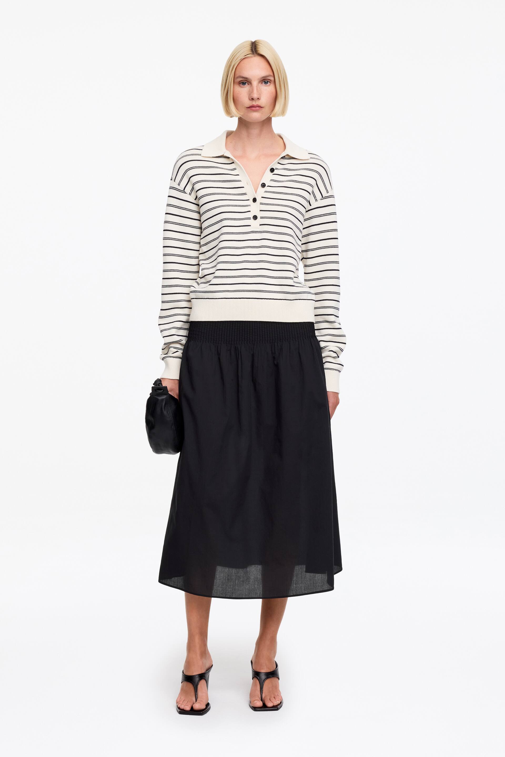 View larger image: A striped polo jumper featuring a cream base with thin black horizontal stripes, a polo collar, and a three-button placket with black buttons. It is designed with long sleeves, ribbed cuffs, and a ribbed hem.