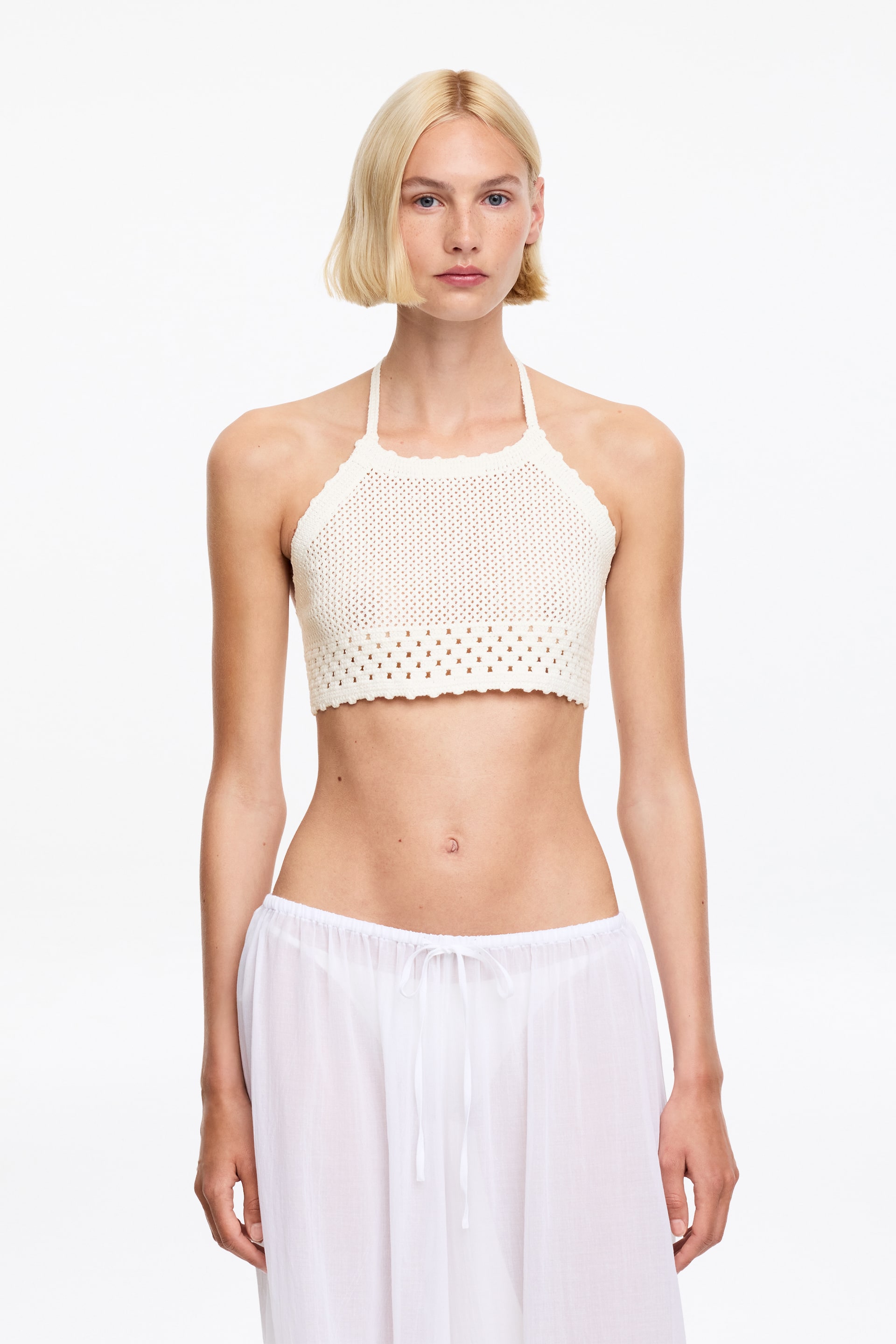Agrandir l'image: Un haut court à col halter en crochet crème présente un décolleté et un ourlet festonnés. Il arbore un motif en tricot ajouré sur l'ensemble du corps principal et s'ajuste étroitement au torse.