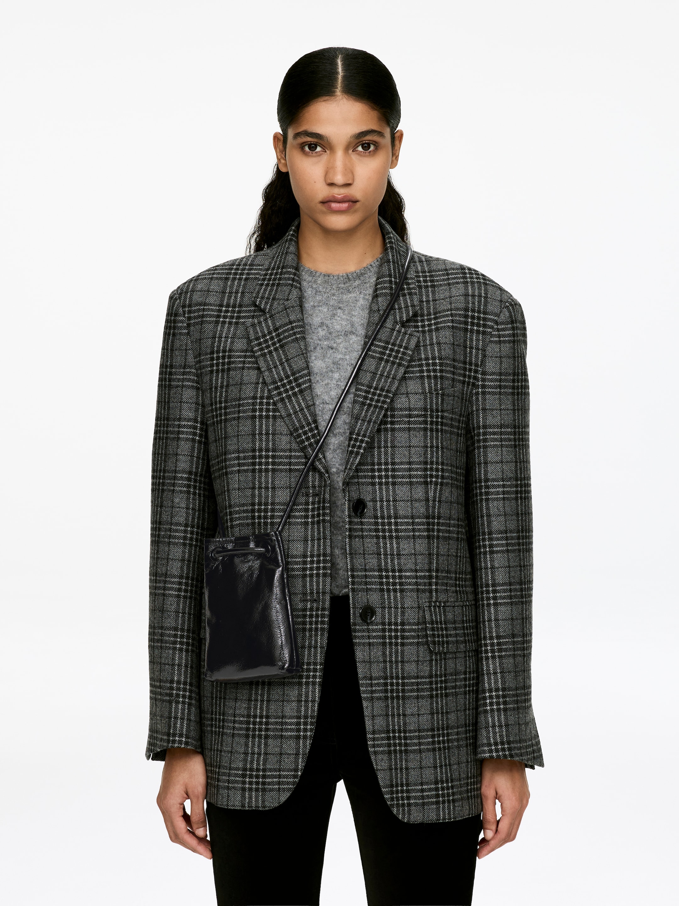 Visa större bild: En mörk läder axelremsväska med en rynkad dragskostängning och tubformade axelremmar bärs av modellen och vilar mot en rutig blazer.