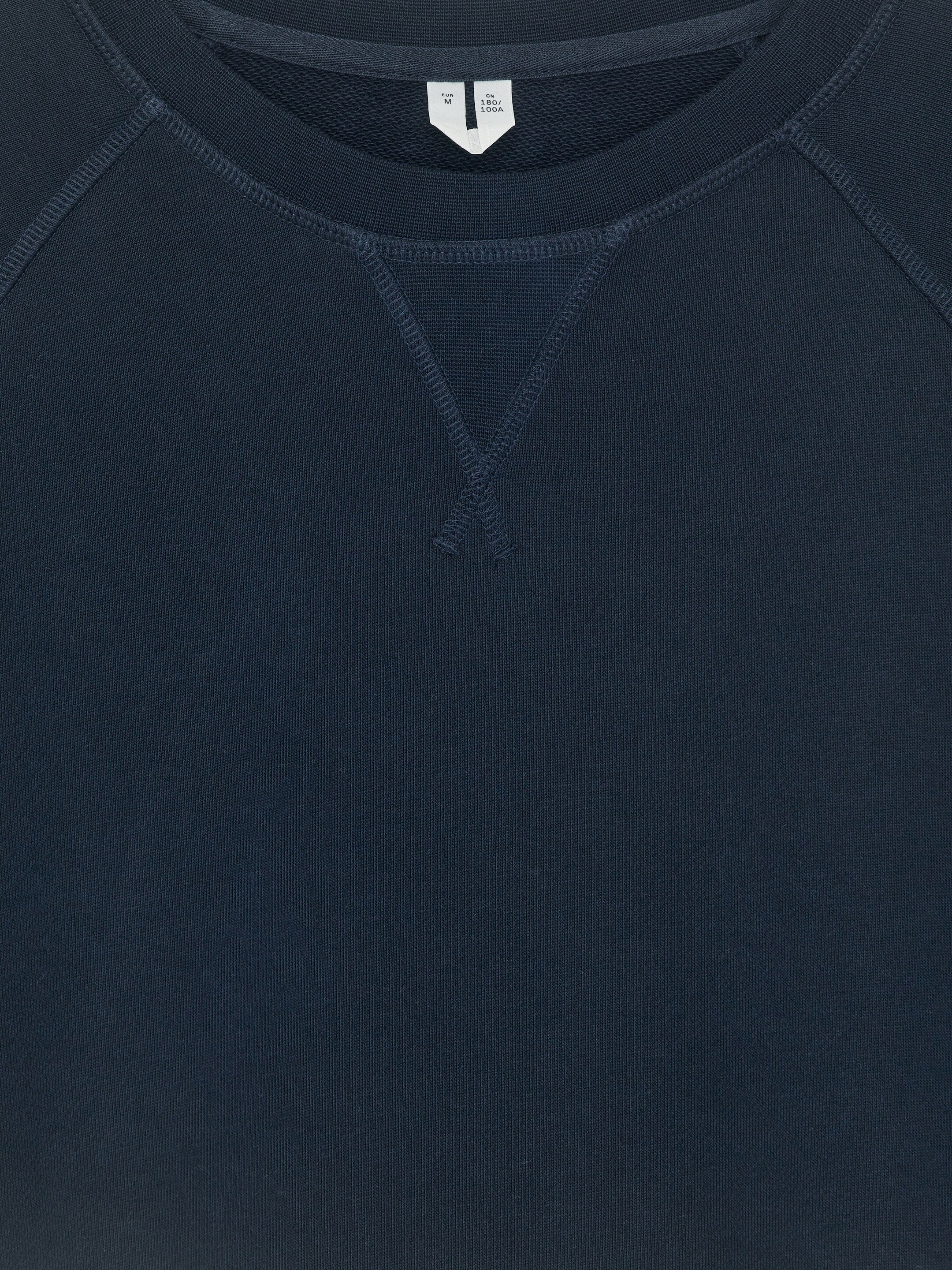 Ingrandisci l'immagine: Una felpa blu scuro con scollo rotondo è mostrata dal davanti, con maniche tagliate a raglan e un colletto a coste. Un dettaglio di cucitura a forma di V è visibile sotto la scollatura.