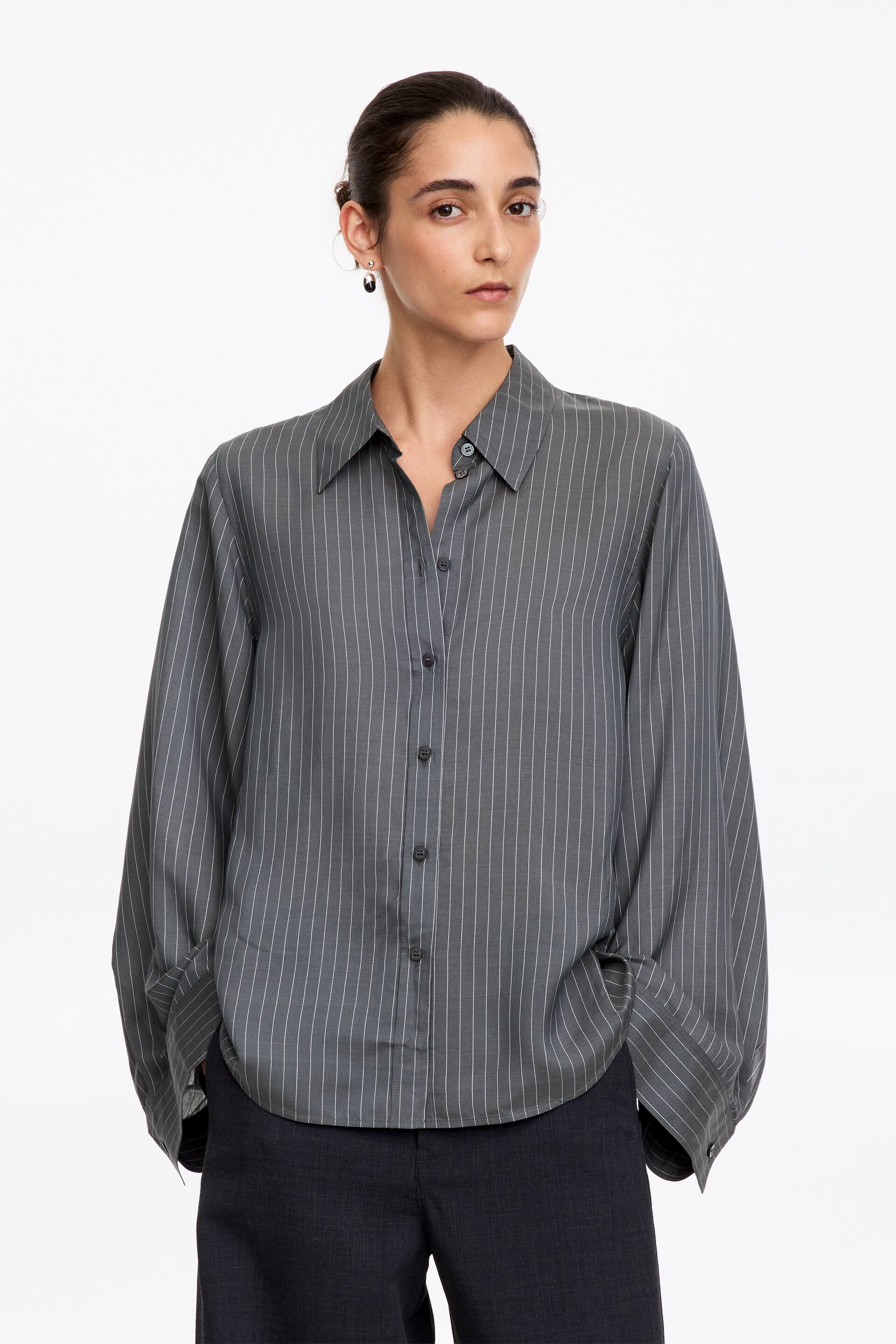 Vis større bilde: En grå pinstripet bluse bæres av modellen og har en avslappet silhuett med vide, flytende ermer og markante mansjetter med knapper. Blusen er stylet med spiss krage og knappestolpe foran, og kombineres med mørke bukser.