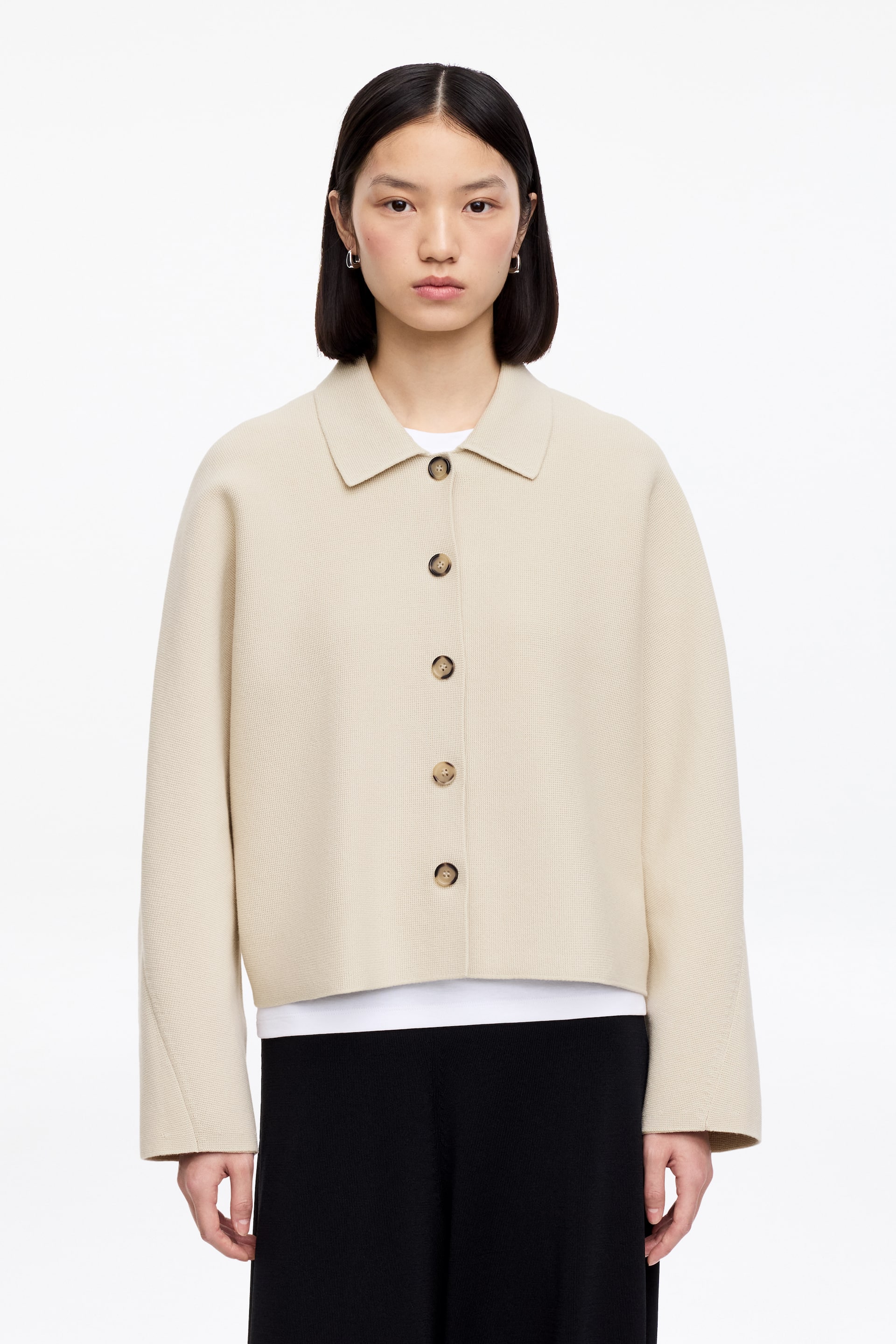 Agrandir l'image: Une veste en tricot crème présente un col pointu, cinq boutons marron clair sur le devant et des manches longues. Elle arbore une longueur raccourcie avec un ourlet droit et est portée sur un t-shirt blanc.