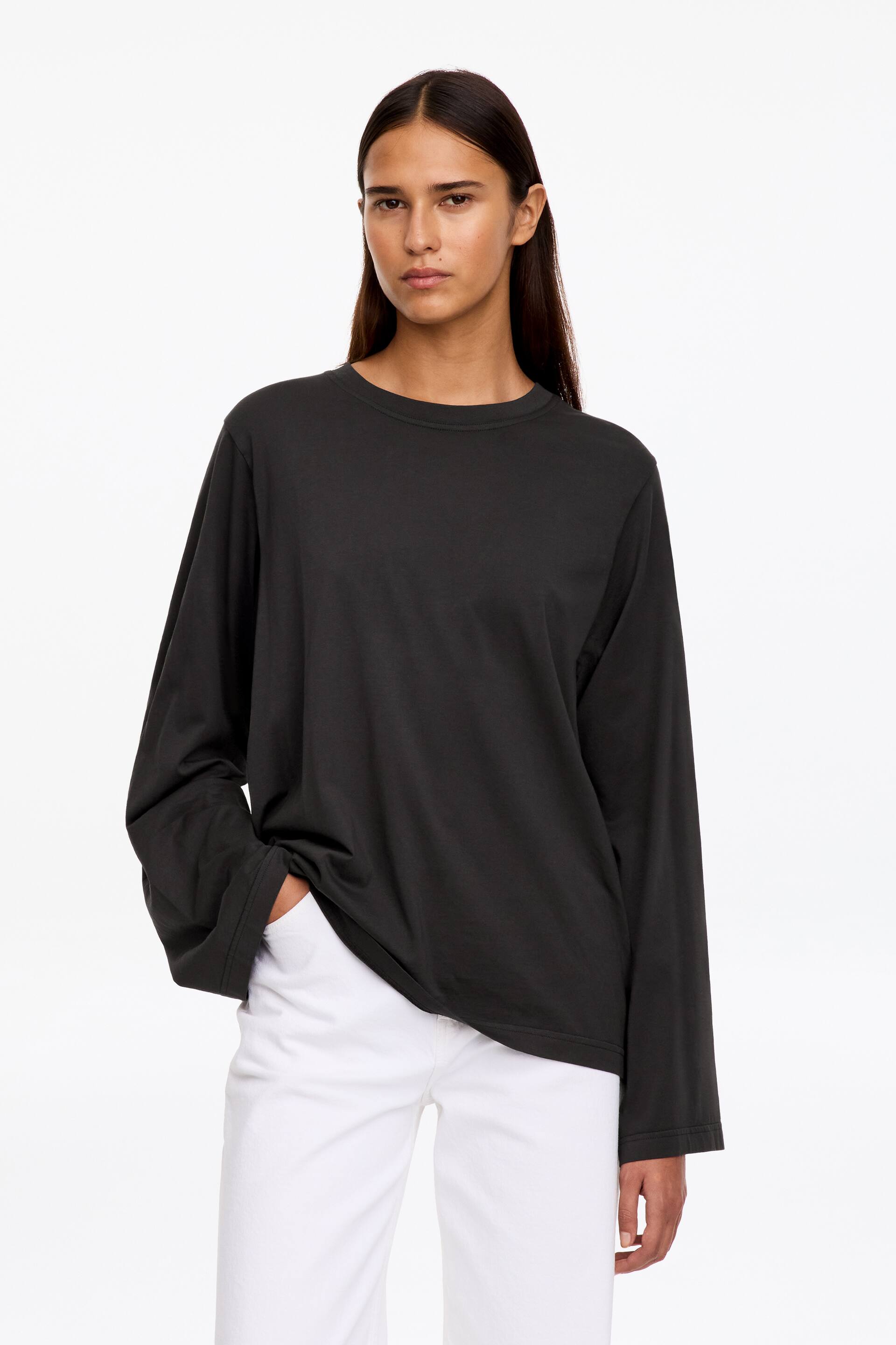 Vezi imaginea mai mare: Un tricou cu mânecă lungă, de culoare gri închis, cu croială lejeră și decolteu rotund, este purtat de model, care stă în picioare cu o mână sprijinită în buzunarul pantalonilor albi. Topul are o siluetă relaxată și mâneci largi.