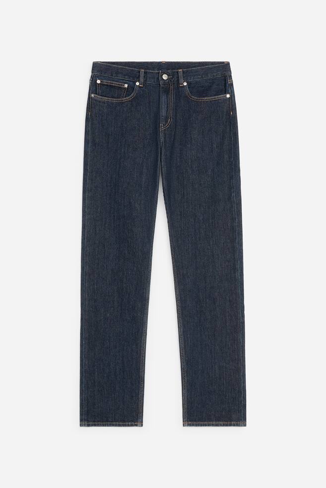 Un par de vaqueros de denim de lavado oscuro aparece de frente, con un diseÃ±o clÃ¡sico de cinco bolsillos con costuras en contraste naranjas y remaches metÃ¡licos. Los vaqueros presentan una silueta de corte recto, una cintura con trabillas para cinturÃ³n y un cierre central de botÃ³n metÃ¡lico Ãºnico. - 1