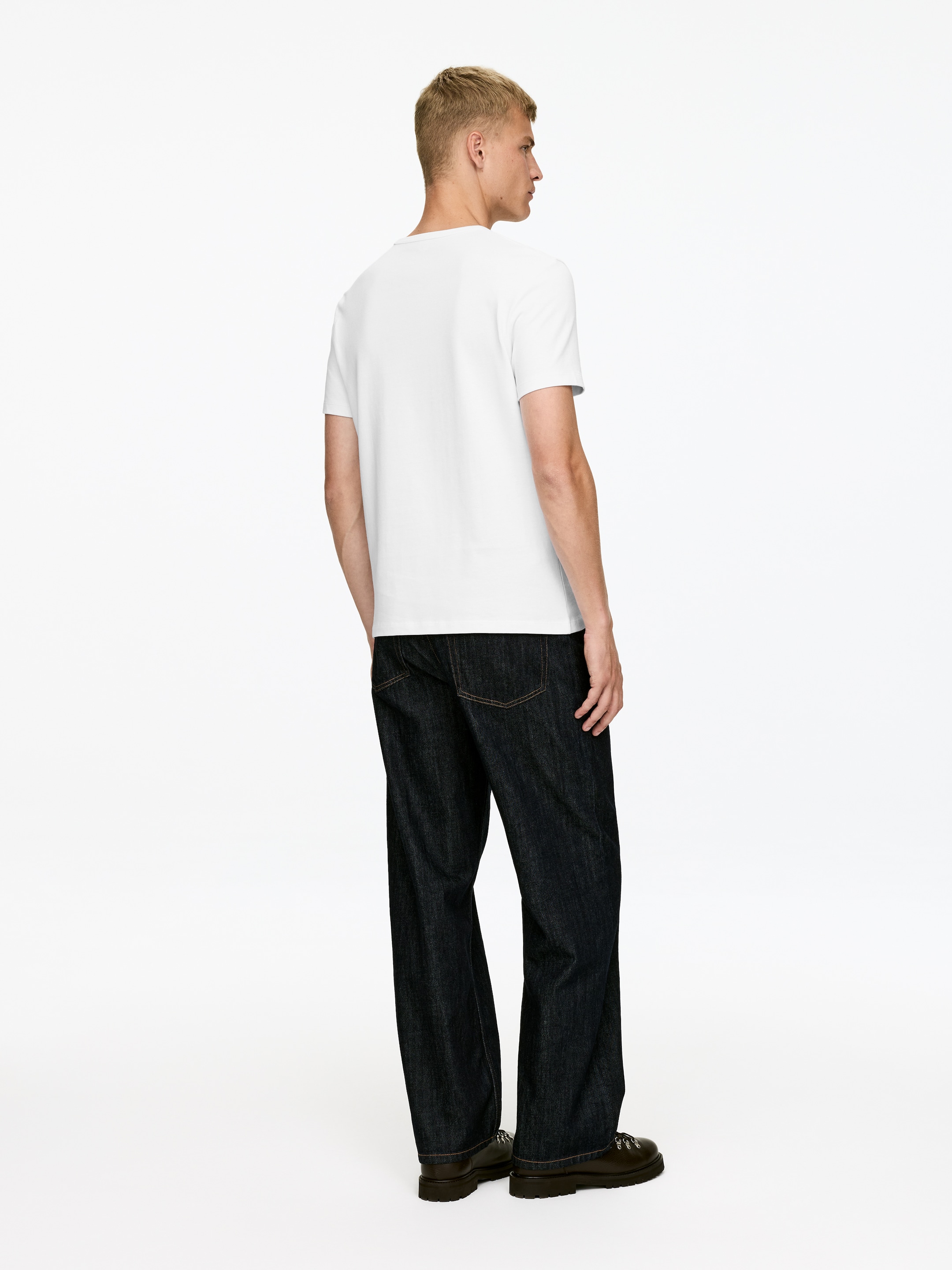 Agrandir l'image: Une personne porte un t-shirt à col rond blanc à manches courtes et un jean large à délavage foncé, présenté de dos en vue 3/4. Le t-shirt est lâche et le jean présente des poches arrière visibles avec des coutures contrastantes.