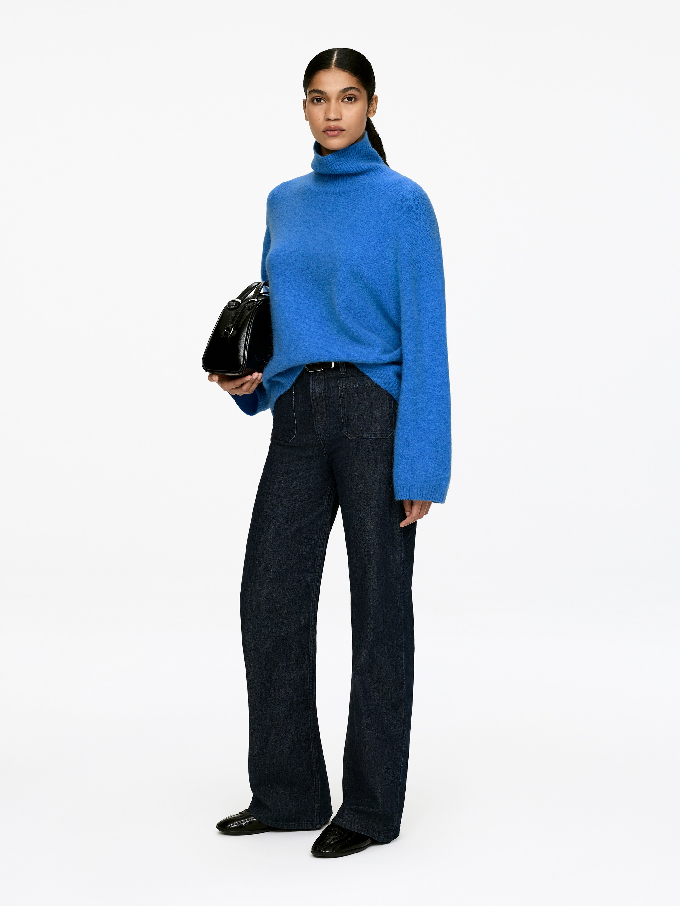 Vis større bilde: En blå turtleneck-genser har en avslappet passform med lange ermer, en litt lodden tekstur, og ribbestrikkede detaljer ved kragen, mansjettene og falden. Mørkvaskede jeans med vide ben har patchlommer foran og er stylet med et svart belte.