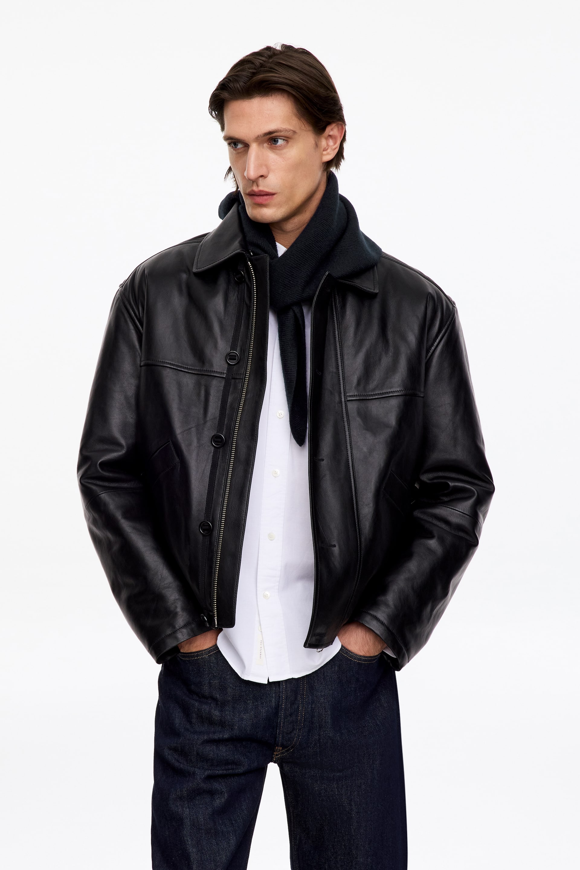 View larger image: A dark, fine-ribbed triangular scarf is being worn wrapped around the model’s neck, layered under an open black leather jacket and over a white button-up shirt.