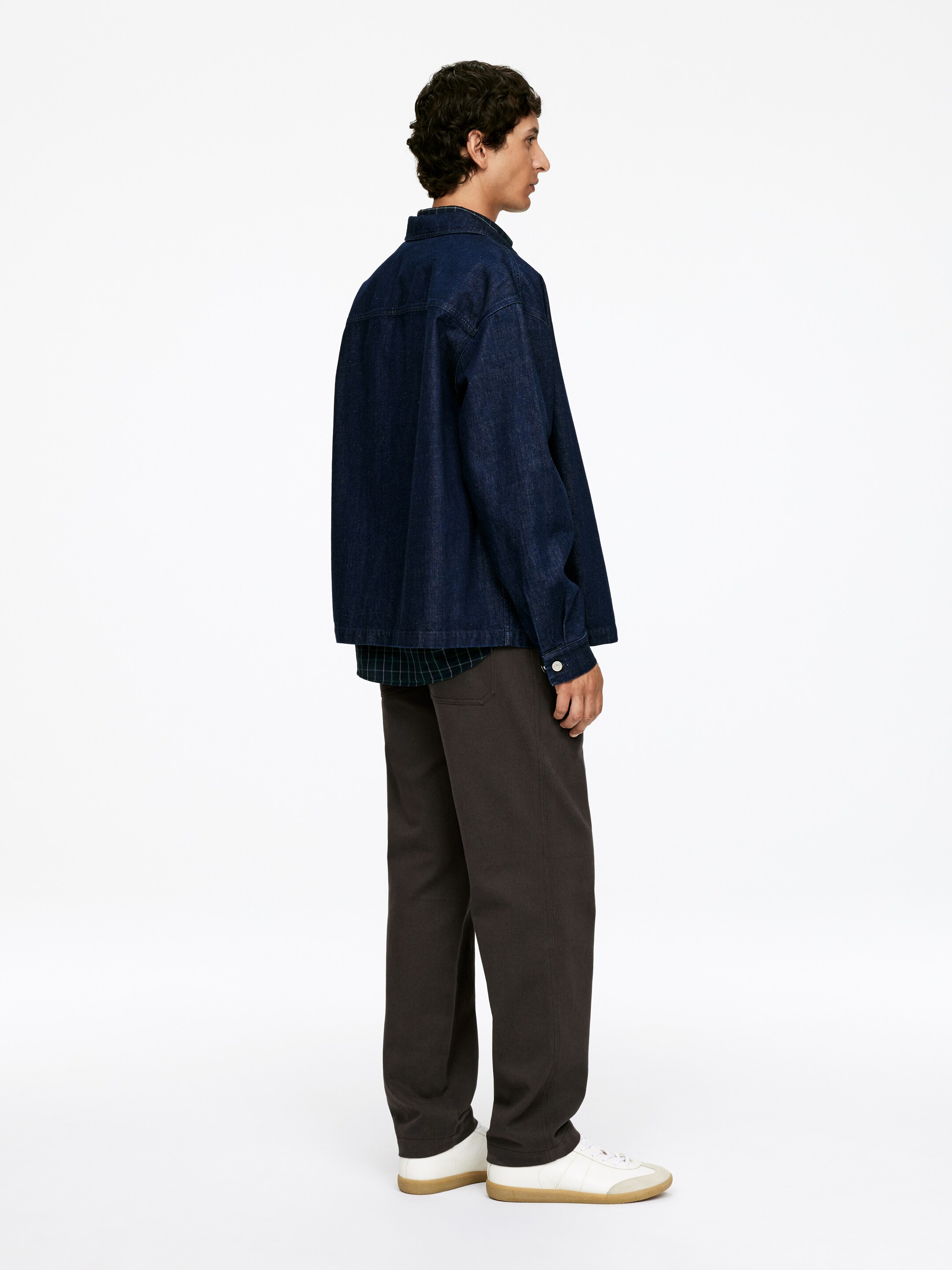 Visualizza immagine più grande: Una sovracamicia in denim scuro, con una vestibilità rilassata, un colletto visibile e polsini con bottoni, è stratificata su una camicia a quadri verde scuro, con il suo orlo che sbuca da sotto. Questo ensemble, che include pantaloni marrone scuro a vestibilità rilassata e sneakers bianche con suole in gomma marrone chiaro, è presentato da una vista di profilo laterale destro, evidenziando la sua silhouette rilassata.