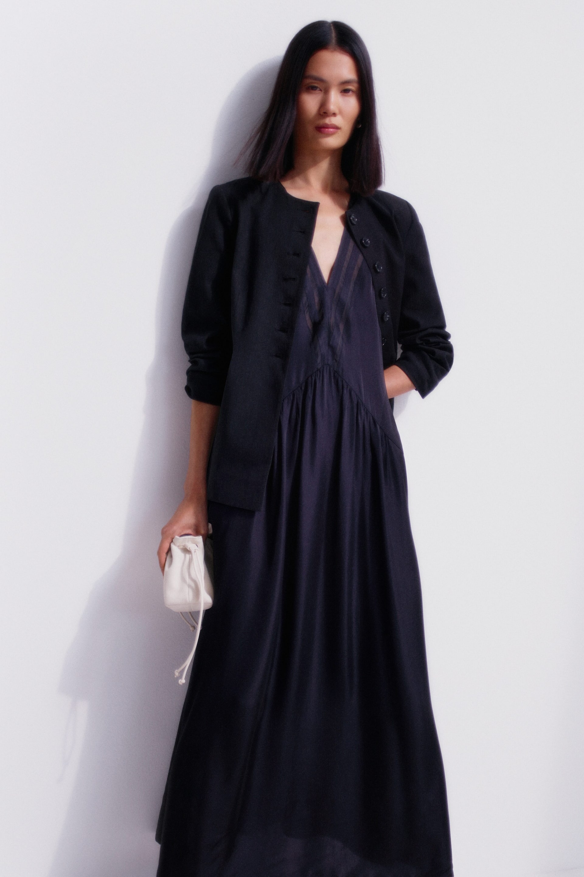 Agrandir l'image: Une veste noire sans col est portée ouverte sur une robe bleu foncé à col en V. La veste présente un encolure arrondie et plusieurs petits boutons sur le devant, tandis que la robe met en avant un corsage froncé avec des rayures verticales subtiles et une jupe longue et fluide.