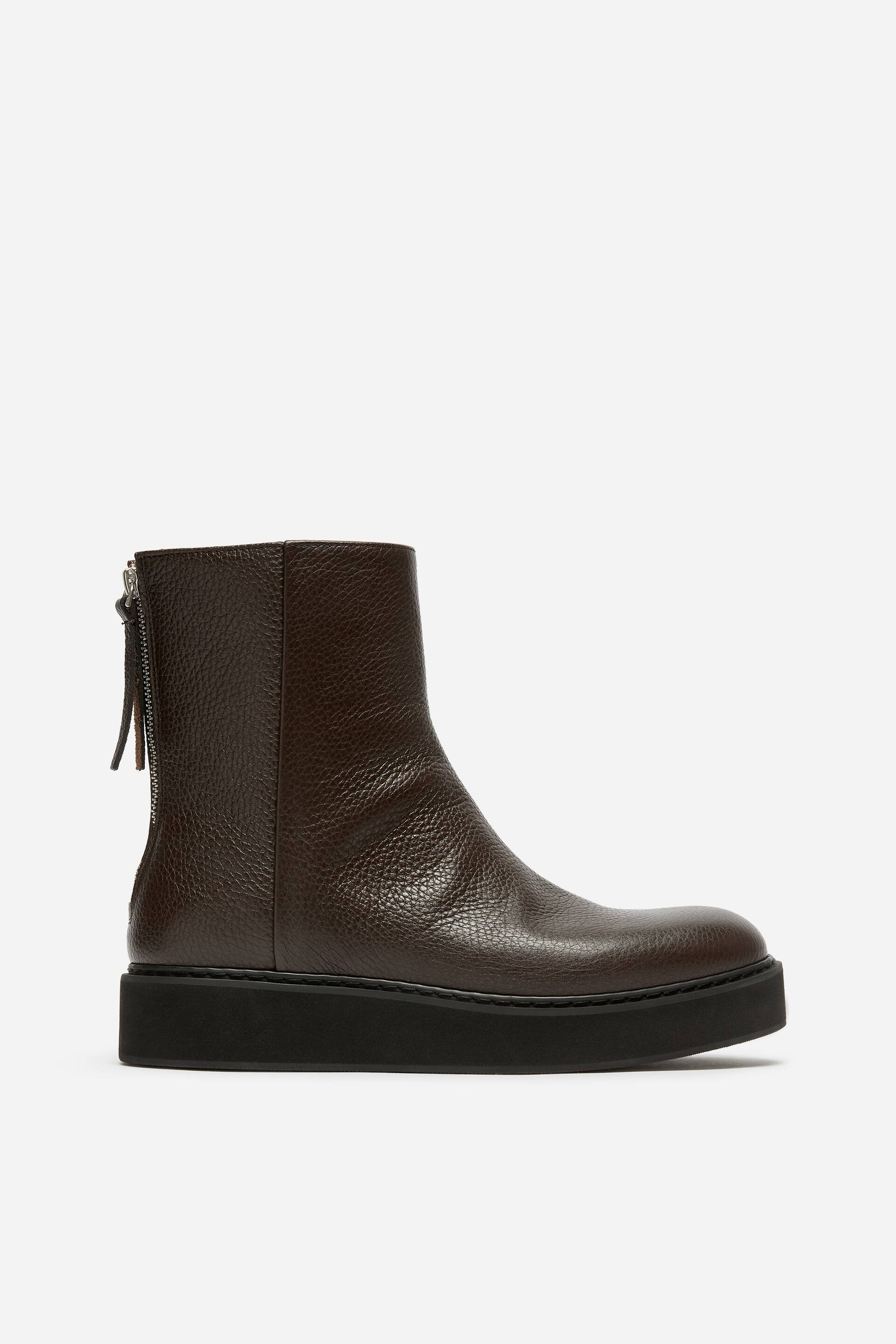 View larger image: A dark brown ankle boot is facing right in side profile, featuring a textured pebbled leather upper and a chunky, flat black platform sole. The design showcases a rounded toe and a visible silver-toned metal zip with a leather pull tab on the back of the shaft.