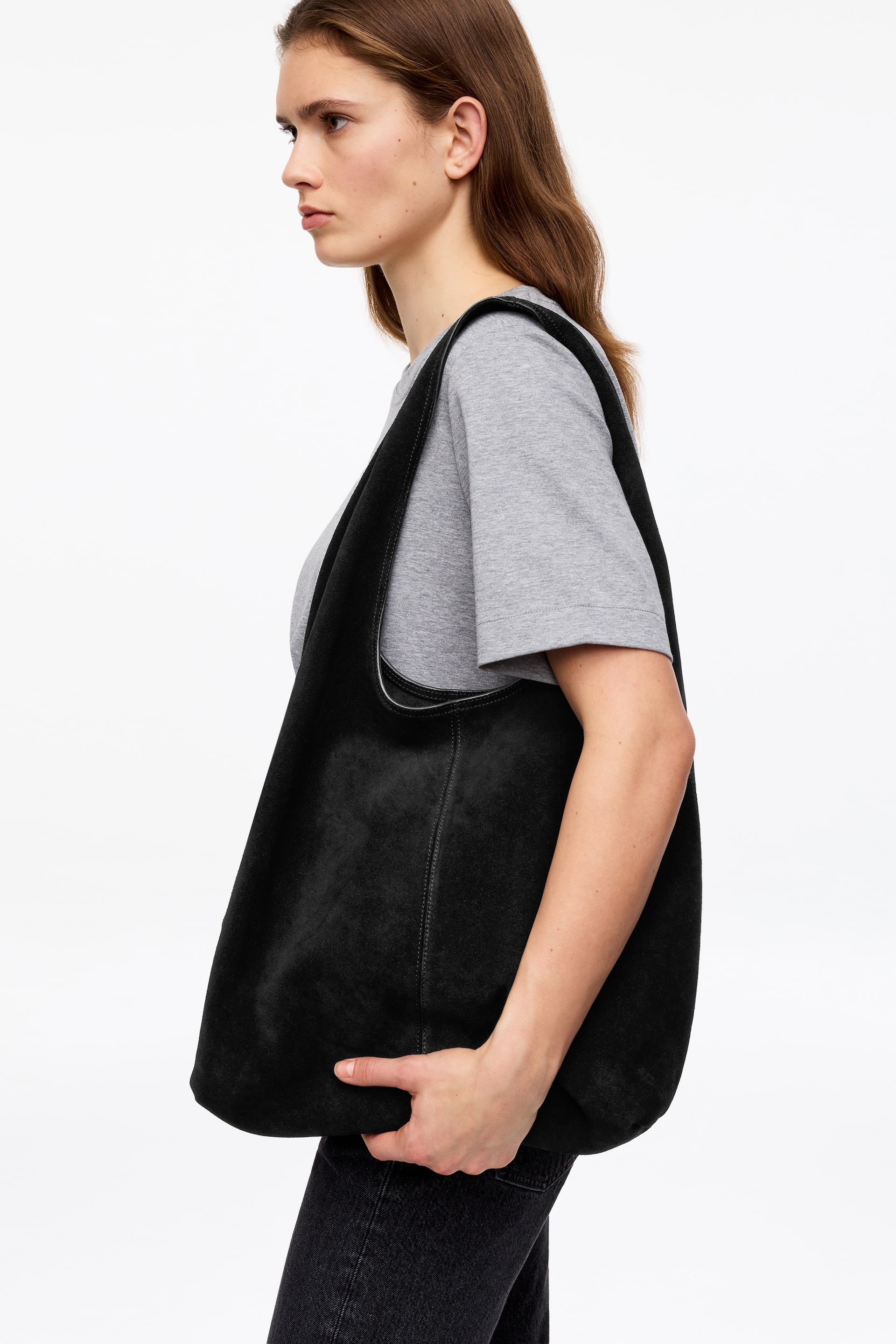 Vis større bilde: En svart semsket slouchy tote bag bæres på høyre skulder, sett fra en sideprofil. Den har en bred, integrert stropp som flyter inn i vesken, og en synlig vertikal søm løper langs siden. Bærerens hånd holder forsiktig på veskens bunn.