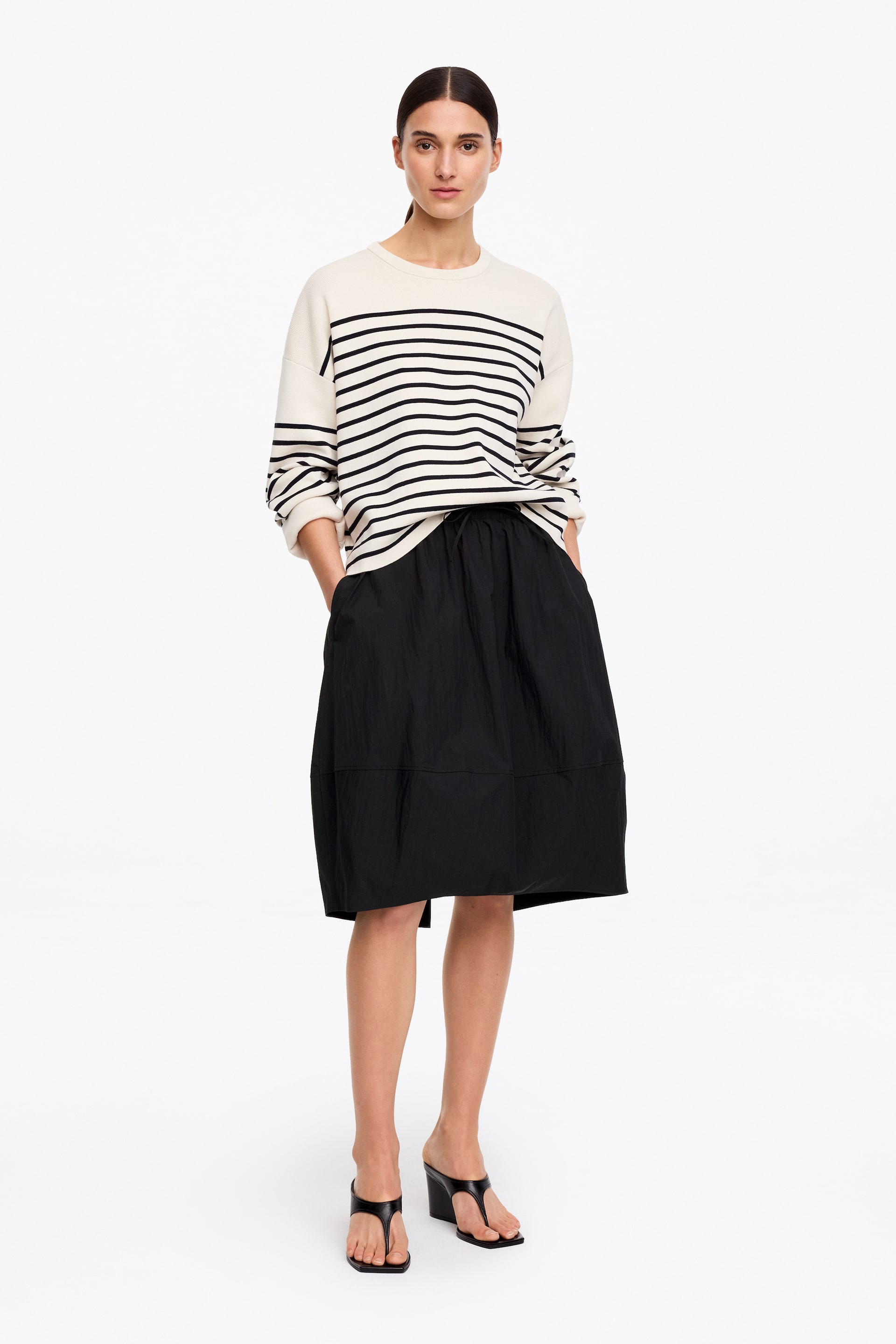 Nagyobb kép megtekintése: "Egy krémszínű, kerek nyakú pulóver fekete vízszintes csíkokkal párosítva egy fekete, középhosszú szoknyával és fekete, telitalpú tangapapucs szandállal. A pulóver laza szabású, míg a szoknya teljes sziluettet mutat be, látható húzózsinóros derékkal."