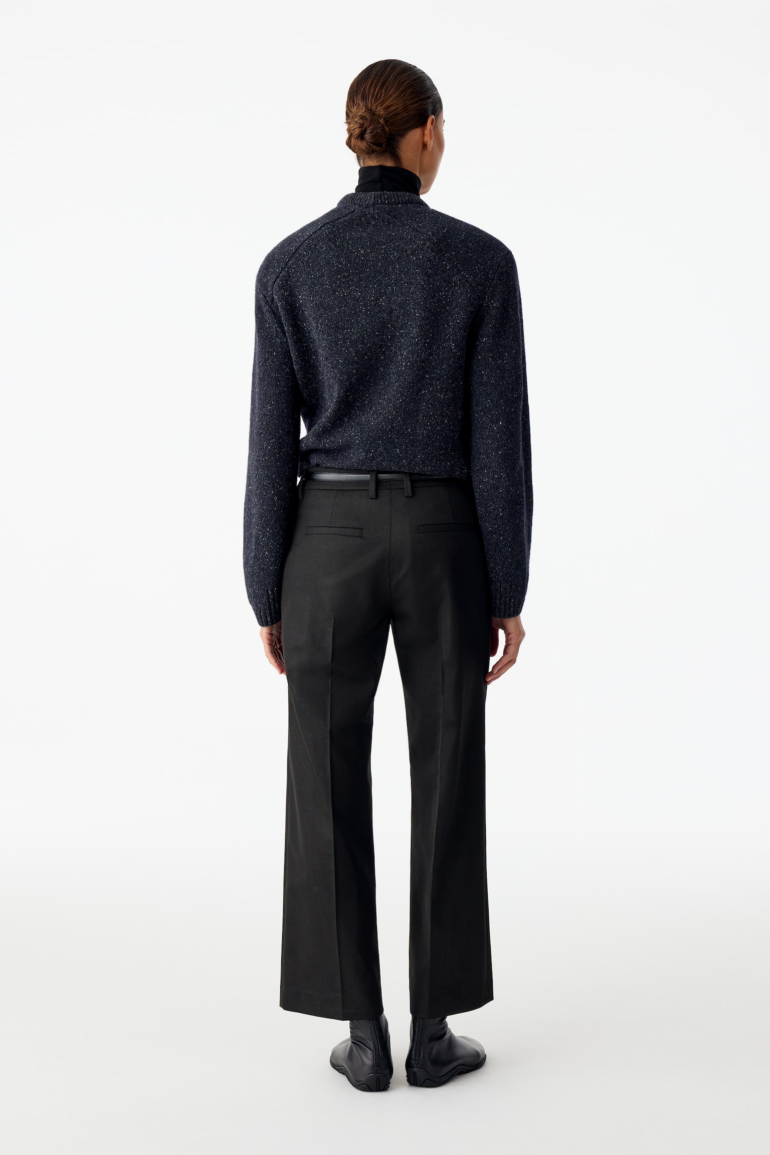 Zwarte pantalon met een recht pijpsilhouet en persvouwen, gedragen door het model en van achteren weergegeven. De enkellange broek heeft achterzakken met paspel en is gestyled met een donkergrijze gebreide trui die ingestopt is en zwarte laarzen. - 1