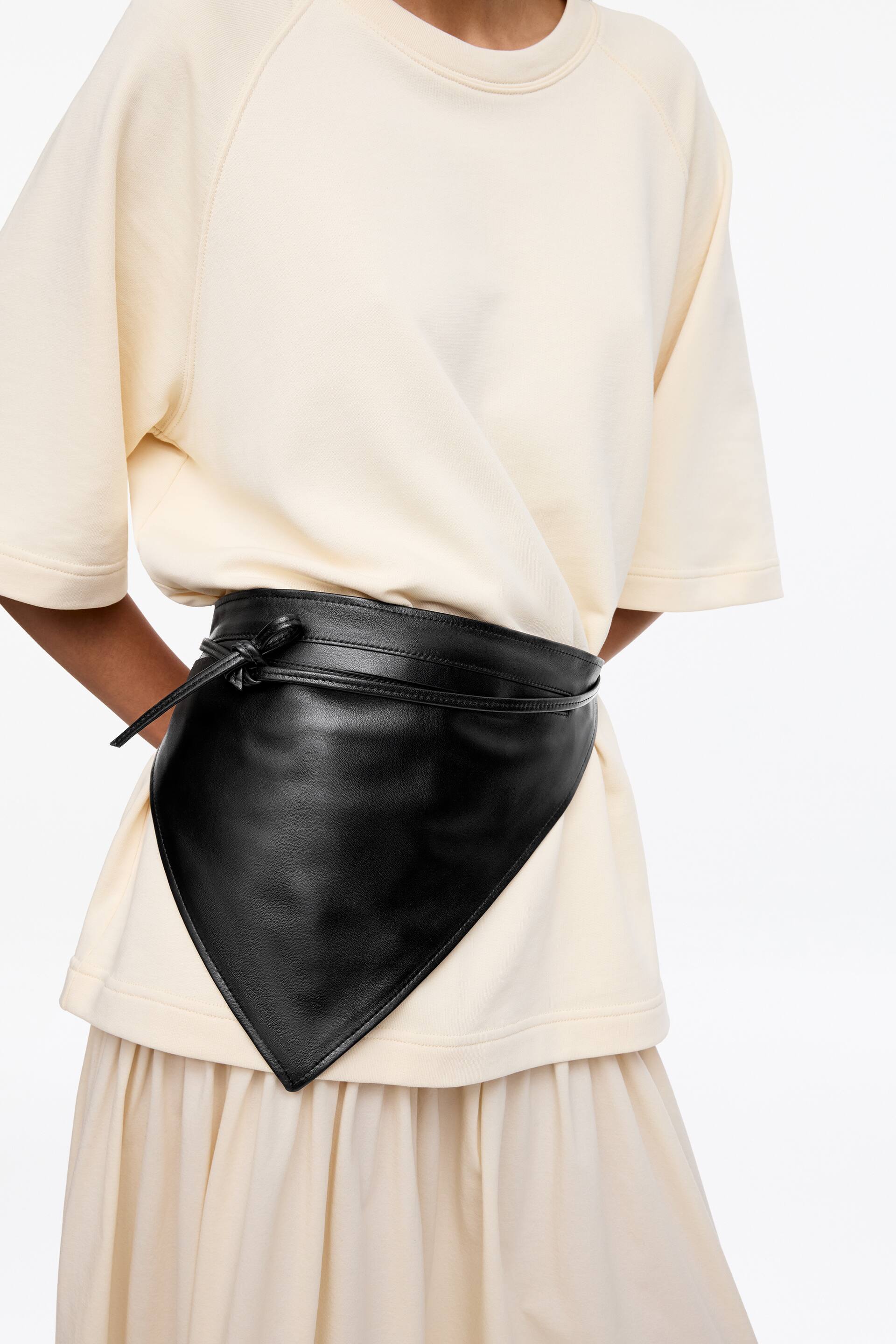 Visa större bild: Ett svart läderomslagsbälte snörar in en krämfärgad topp och lager på lager över en krämfärgad kjol. Bältet har en bred design med en spetsig, triangulär framsida och knyts med tunna band på vänster sida.