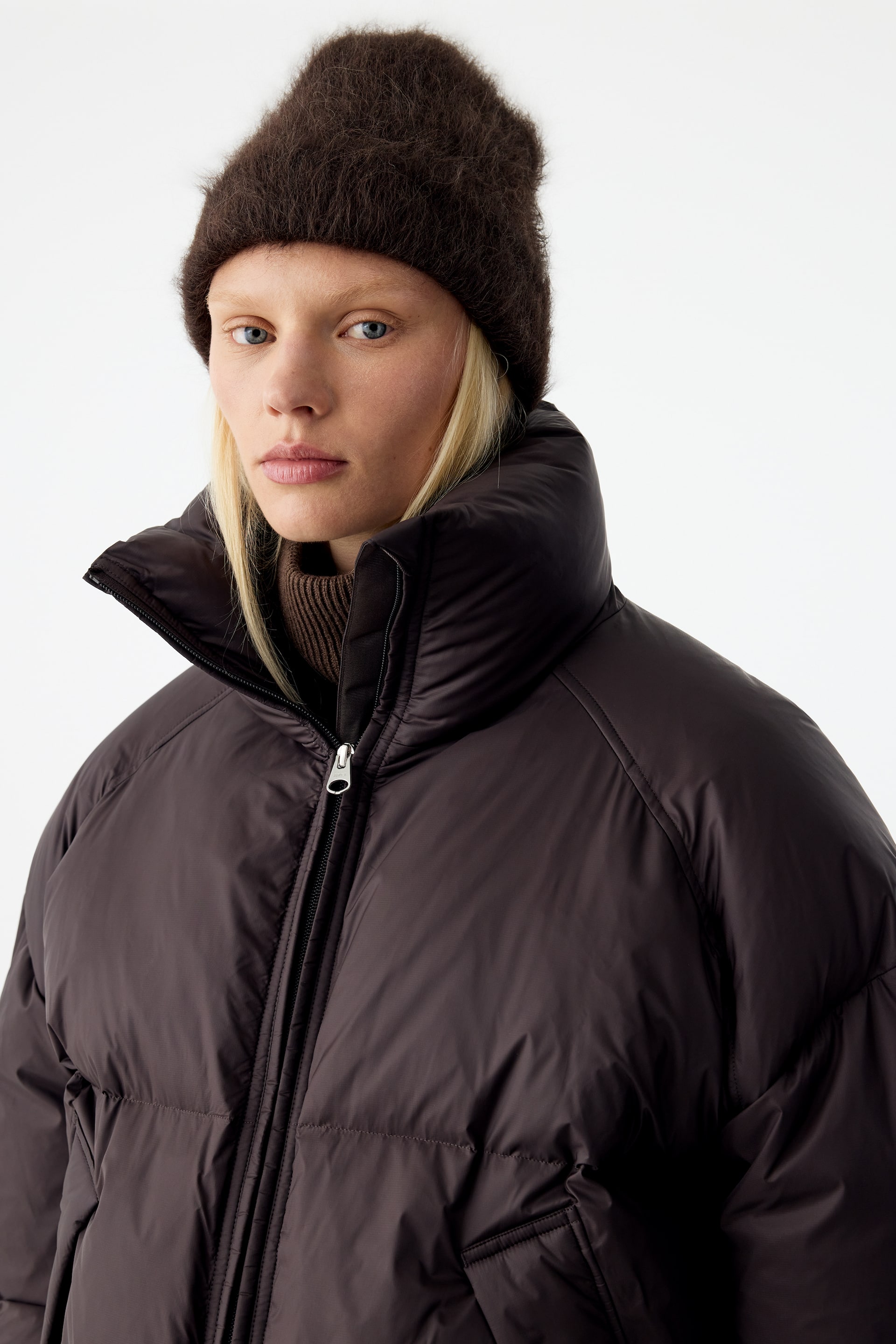 View larger image: A dark brown, fluffy knitted beanie is being worn by the model, pulled low over the forehead. The brushed-texture beanie is styled with a matching dark brown puffer jacket and a high-neck ribbed sweater.