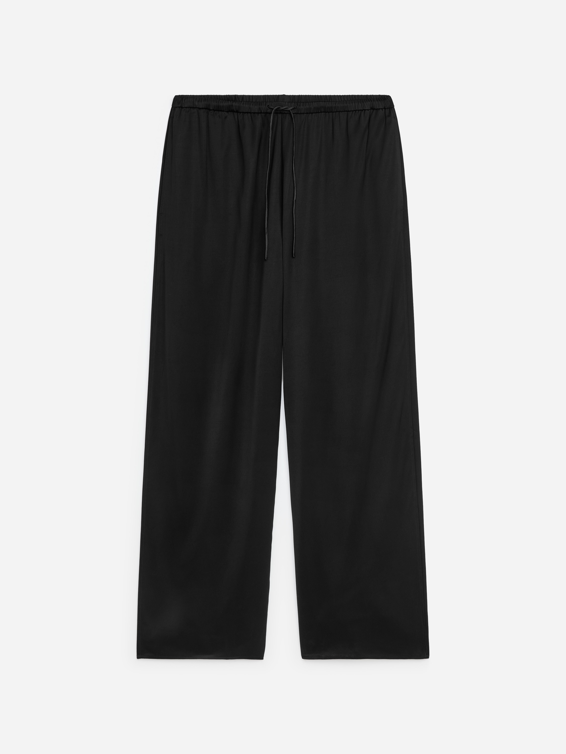 Pantaloni satinati neri dal taglio rilassato, caratterizzati da una silhouette fluida e da una vita con coulisse, indossati dalla modella, che è in piedi con le mani inserite nelle tasche laterali. I pantaloni sono abbinati a un maglione in maglia a V grigio scuro, sovrapposto a una T-shirt bianca.