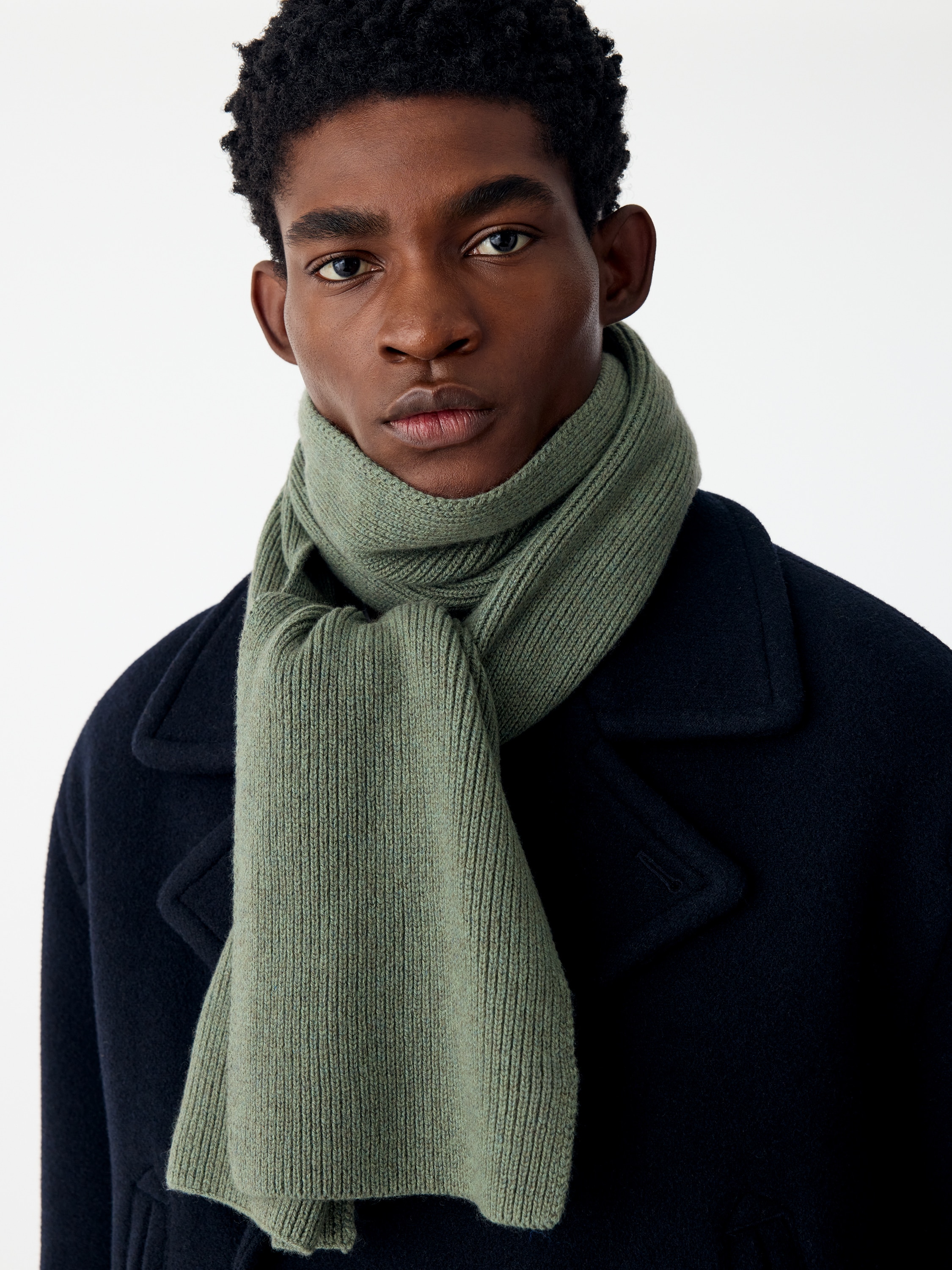 A sage green knitted scarf is shown from the front, folded vertically to showcase its textured, ribbed design. The scarf features a consistent vertical rib-knit pattern with clean, straight edges.