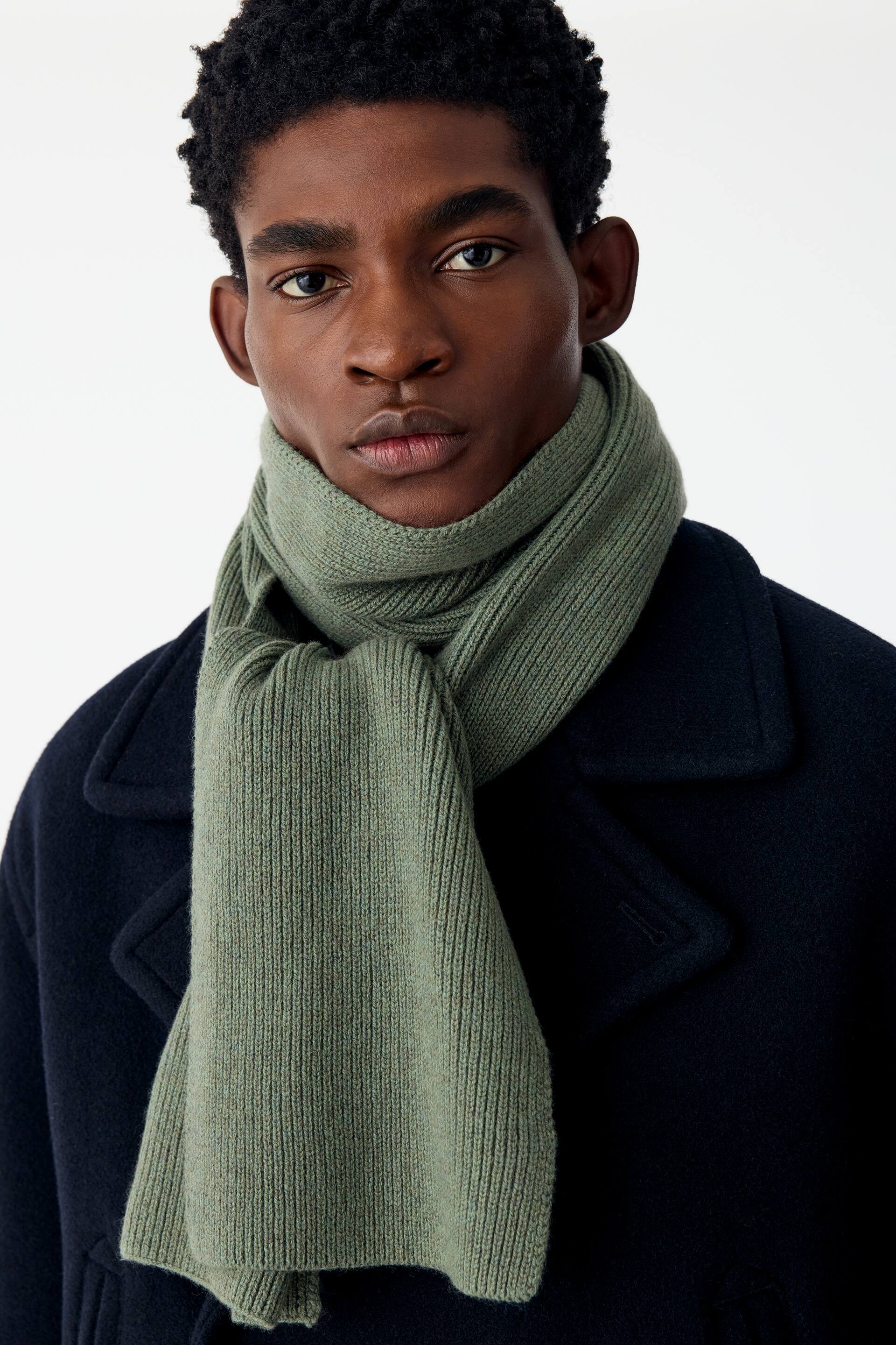 View larger image: A sage green rib-knitted scarf is being worn wrapped around the neck of the model, layered over a dark navy coat. The scarf features a fine, compact ribbed texture, with one end draping down the front of the coat.