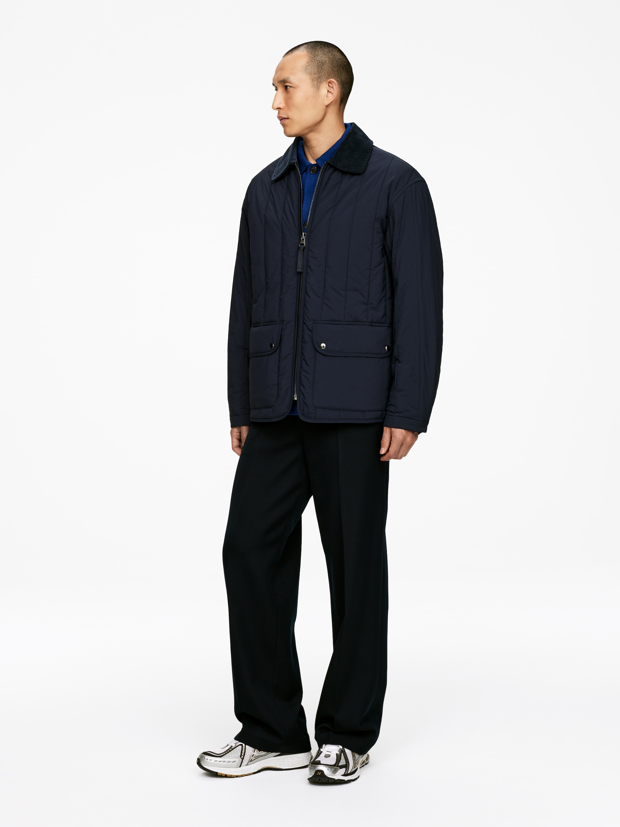 Grösseres Bild ansehen: Eine marineblaue, gesteppte Jacke wird aus einer Dreiviertel-Front- und Seitenansicht präsentiert, mit vertikaler Steppung, einem dunklen Cordkragen und einem silberfarbenen Reißverschluss in voller Länge. Zwei große aufgesetzte Taschen mit Klappen- und Druckknopfverschlüssen sind auf der Vorderseite sichtbar, darunter ist ein blaues Poloshirt zu sehen.